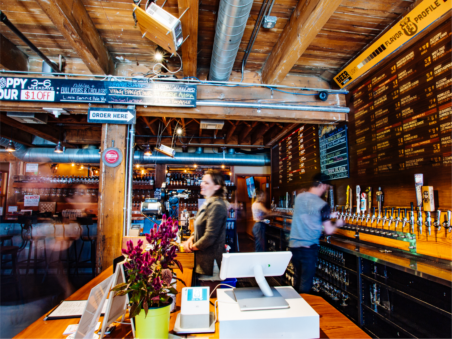 Bartenders at a brewery taproom counter beneath a chalkboard beer menu