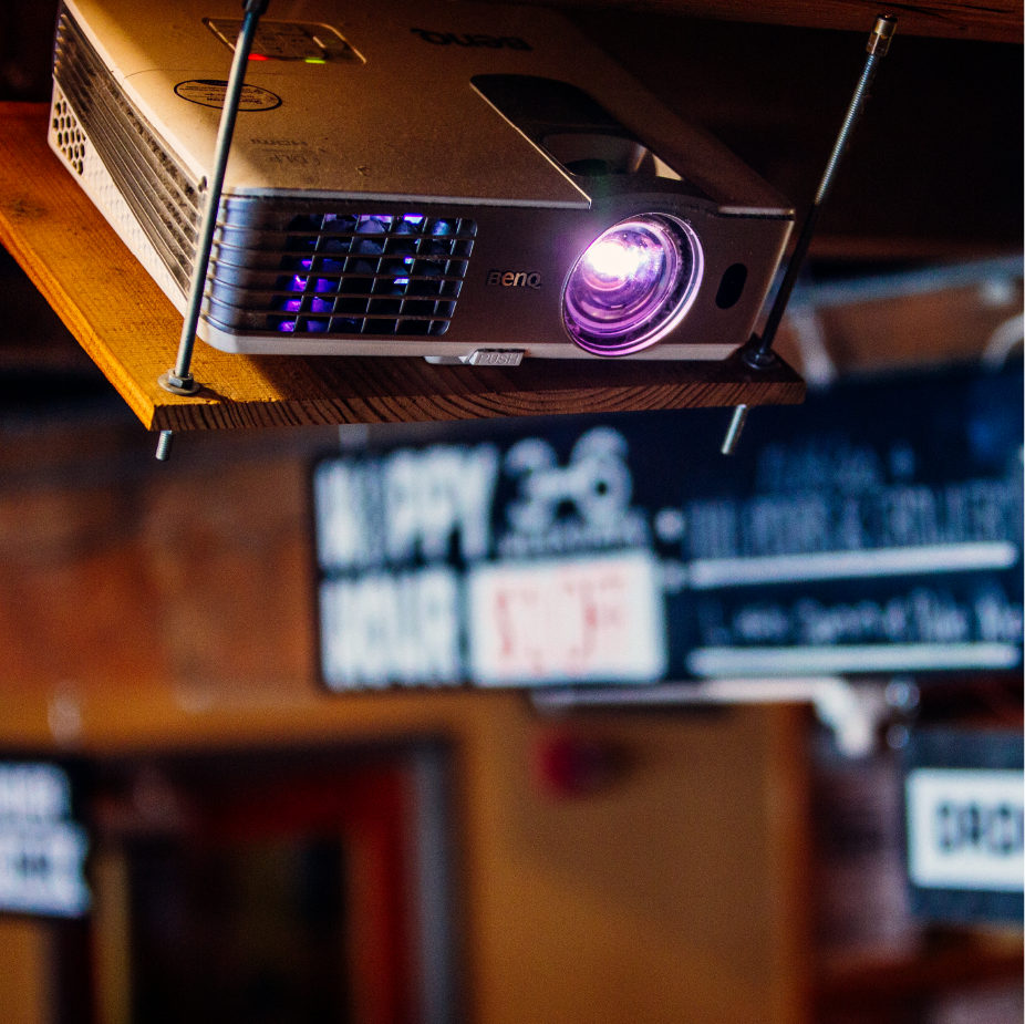 BenQ projector mounted on a wood shelf glowing purple above blurred Tap and Growler signs