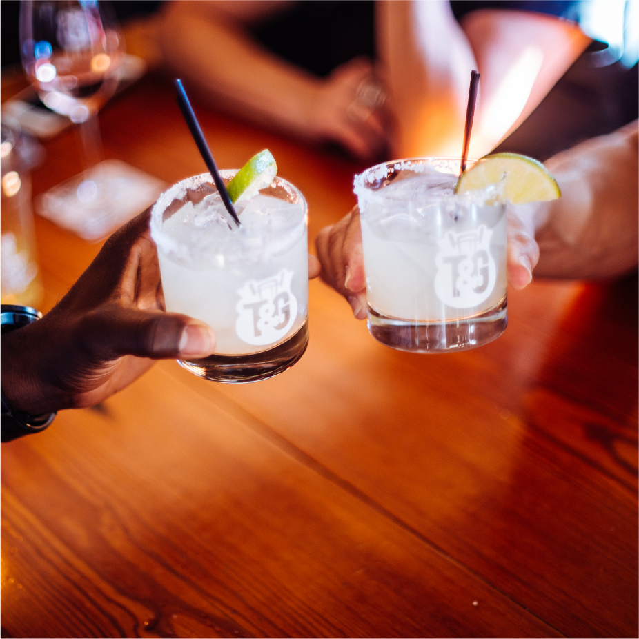Two hands toasting etched cocktail glasses of margaritas with lime over a wooden table