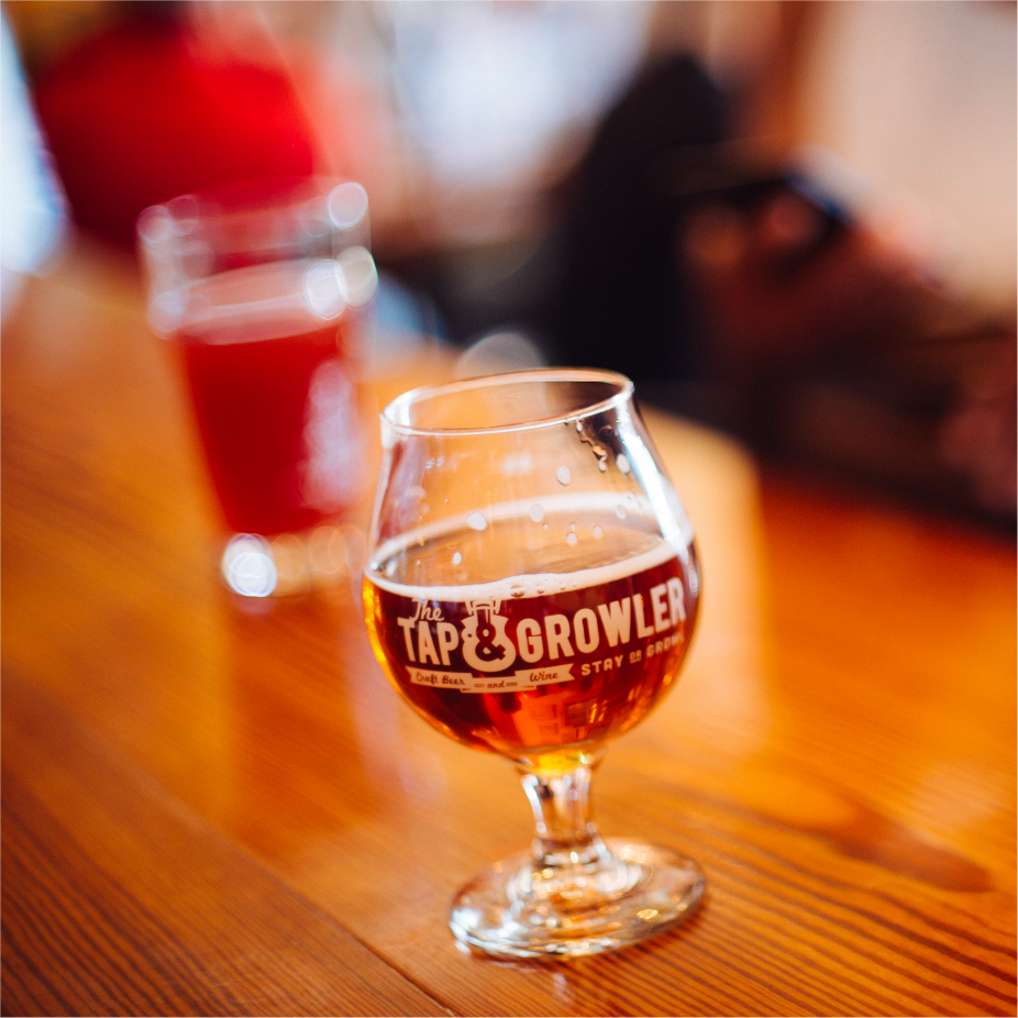 Glass etched with The Tap and Growler logo holding amber beer on a wooden bar