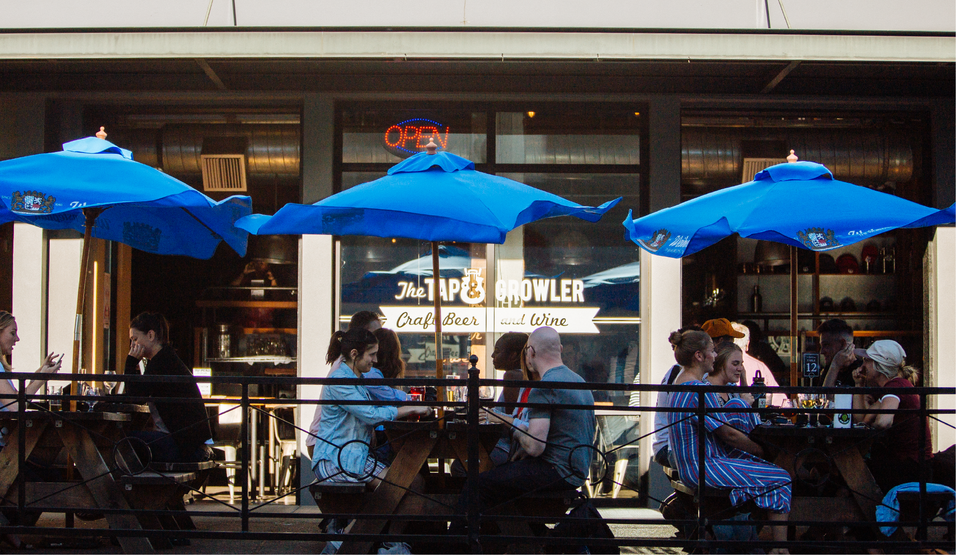 Diners under blue umbrellas at The Tap & Growler patio, Craft Beer and Wine, Open sign in window