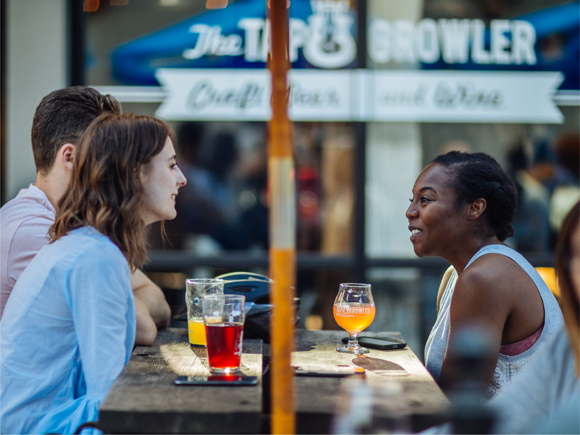 Three friends talking over beers at an outdoor table outside The Tap and Growler