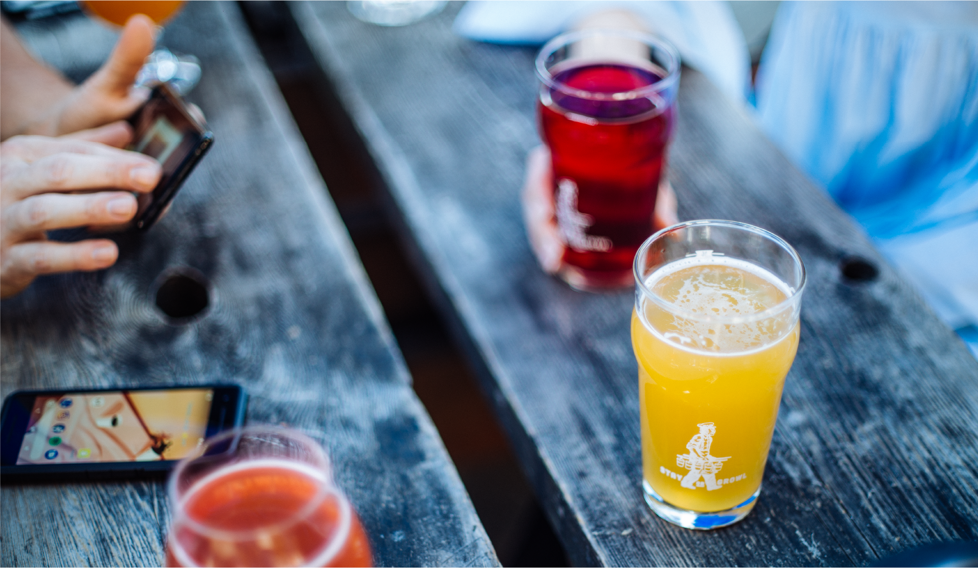 Glasses of red and hazy golden beer on a weathered picnic table, phones nearby