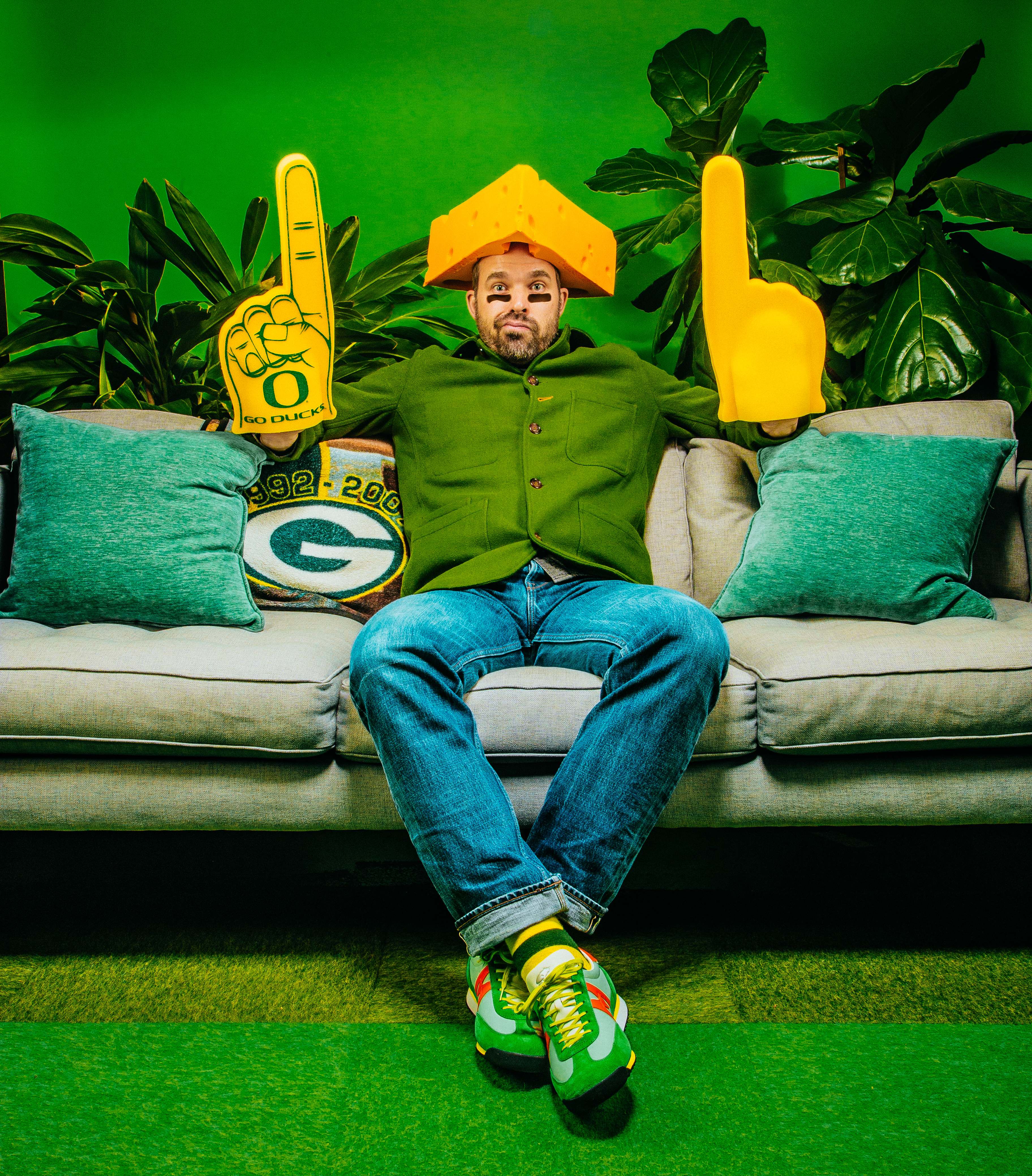 Man on a couch wearing a cheesehead hat, holding Oregon Ducks and foam fan fingers against a green backdrop