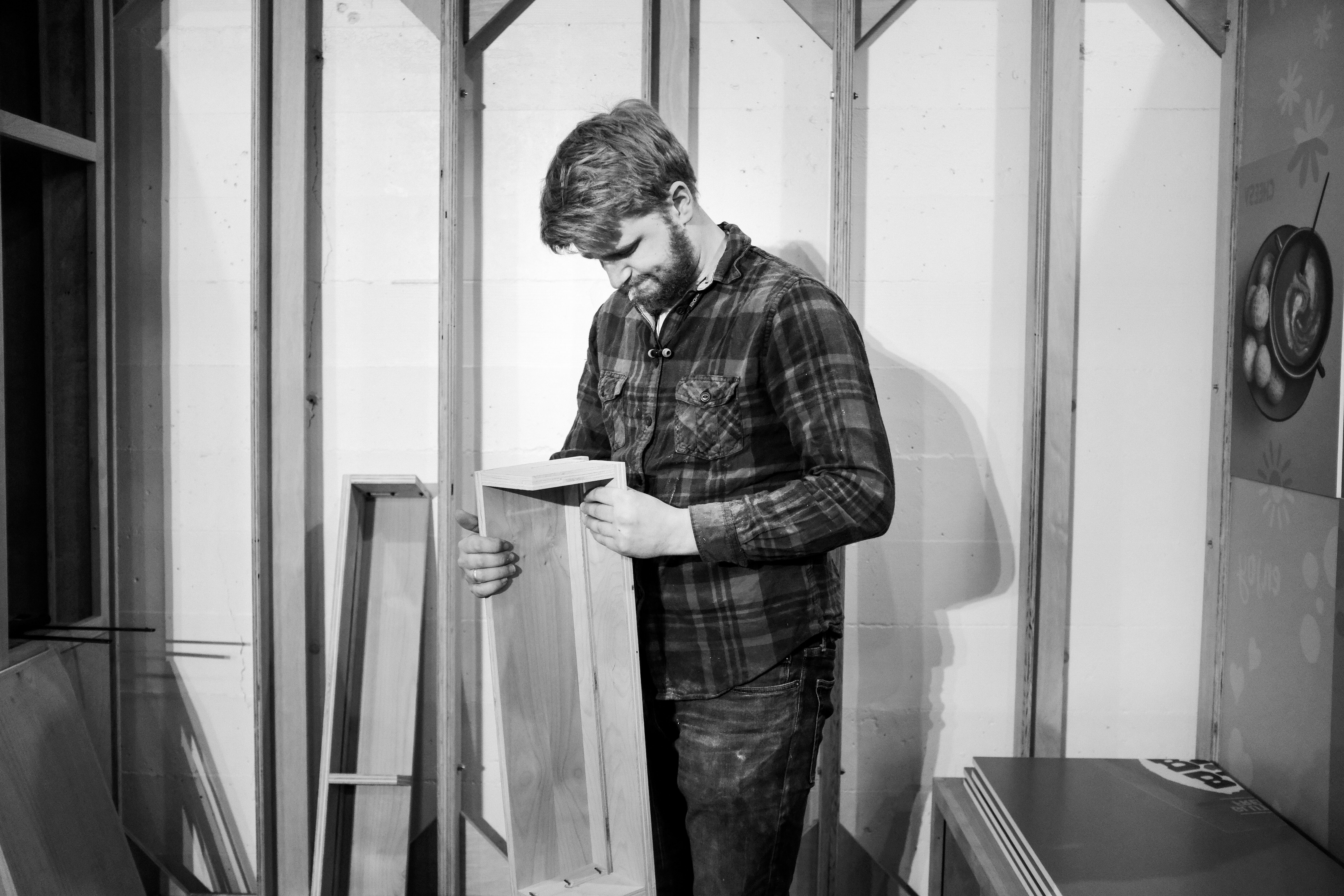 Man in plaid shirt assembling a wooden booth frame in the Drawn workshop, black and white