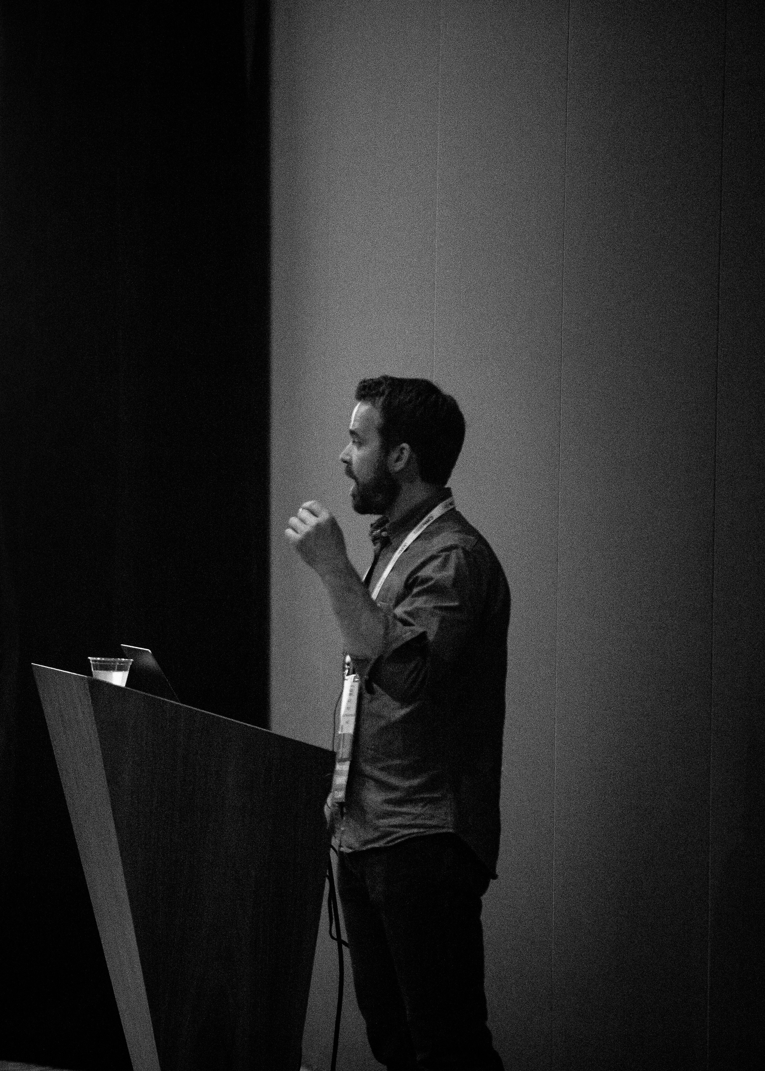 Bearded man speaking and gesturing at a lectern on a dim conference stage, black and white