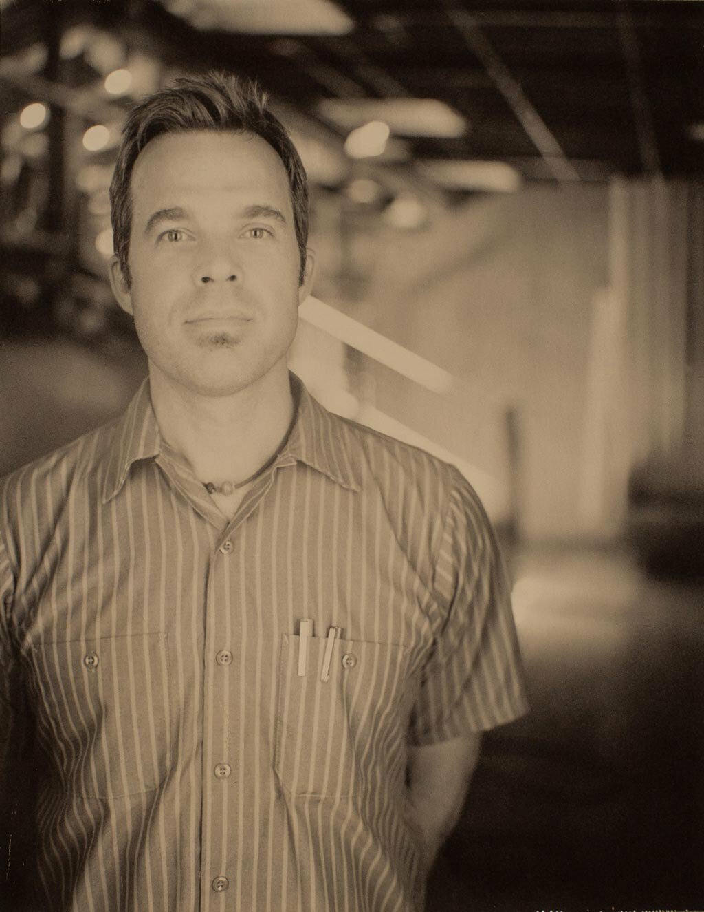 Sepia-toned portrait of a man in a striped short-sleeve shirt standing in an open workspace