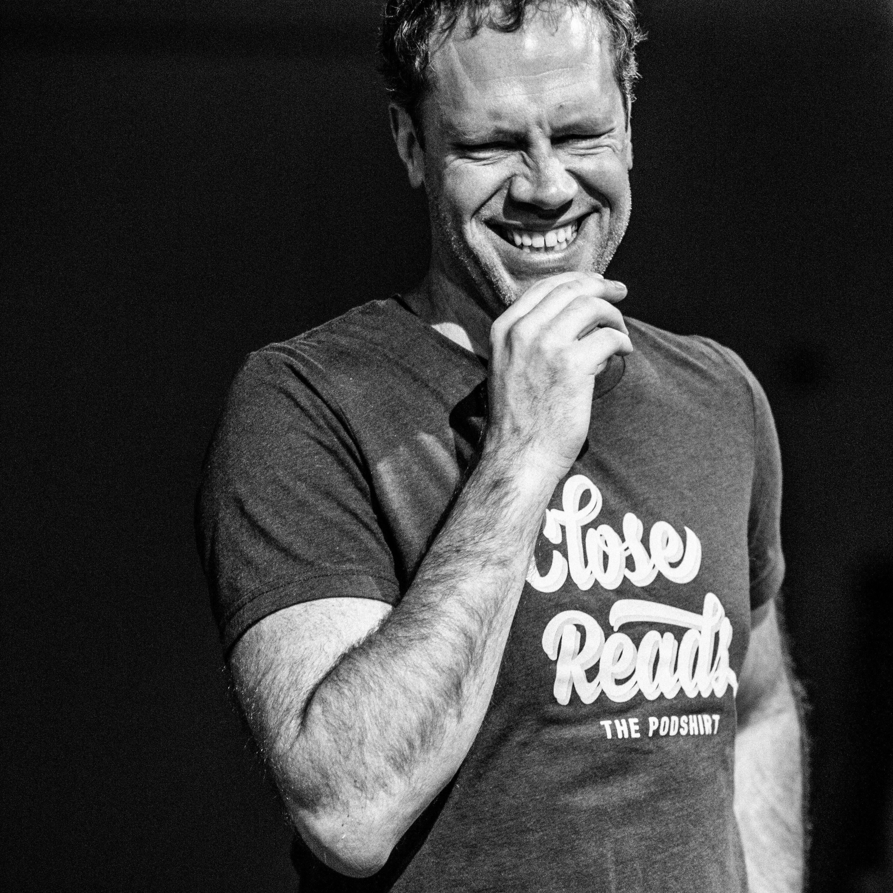 Laughing man holding a microphone, wearing a Close Reads The Podshirt tee, black and white