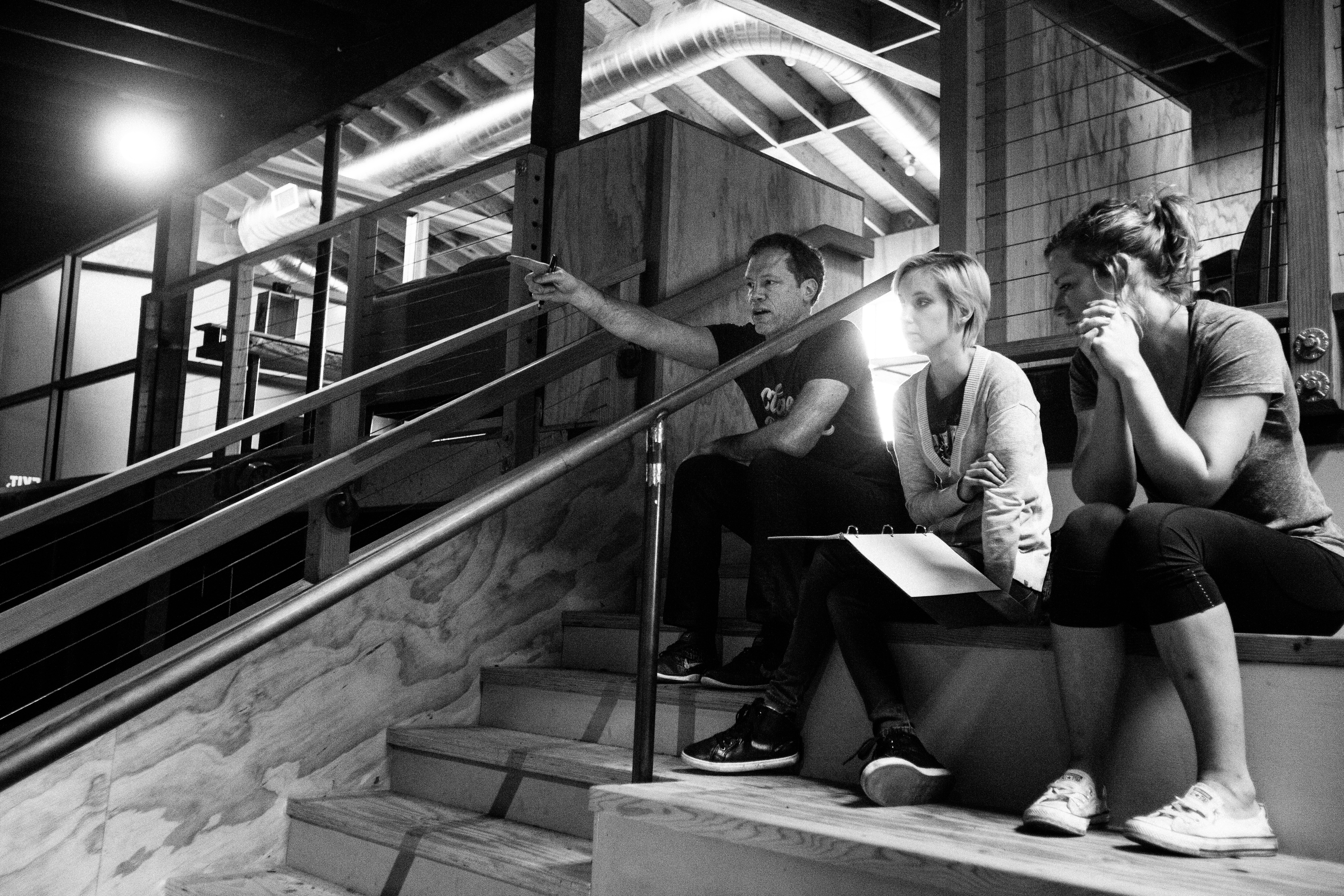 Three people seated on stairs reviewing work as one points, in a dim wood interior, black and white