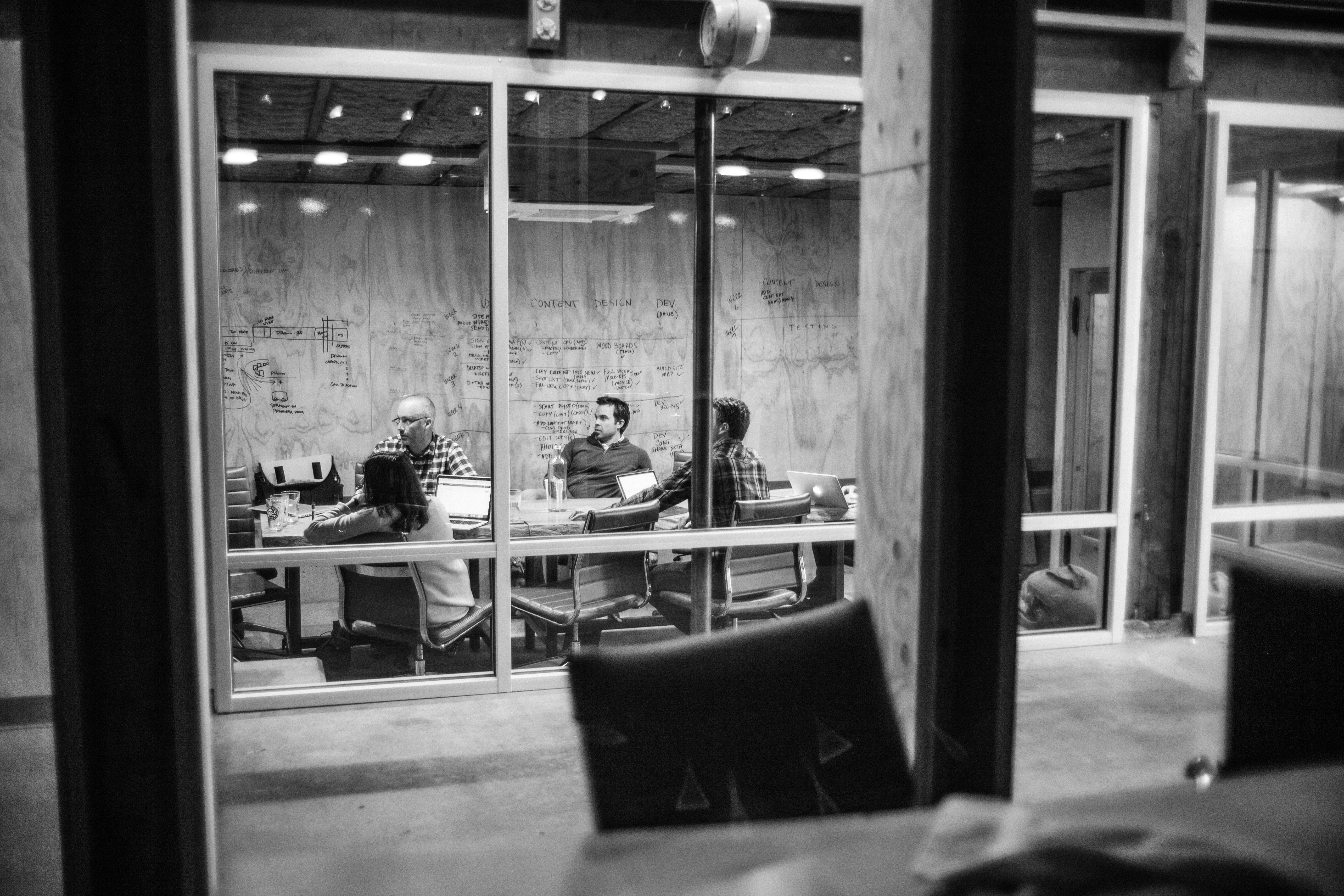 View through a glass wall of a team meeting in a wood-paneled Drawn conference room, black and white