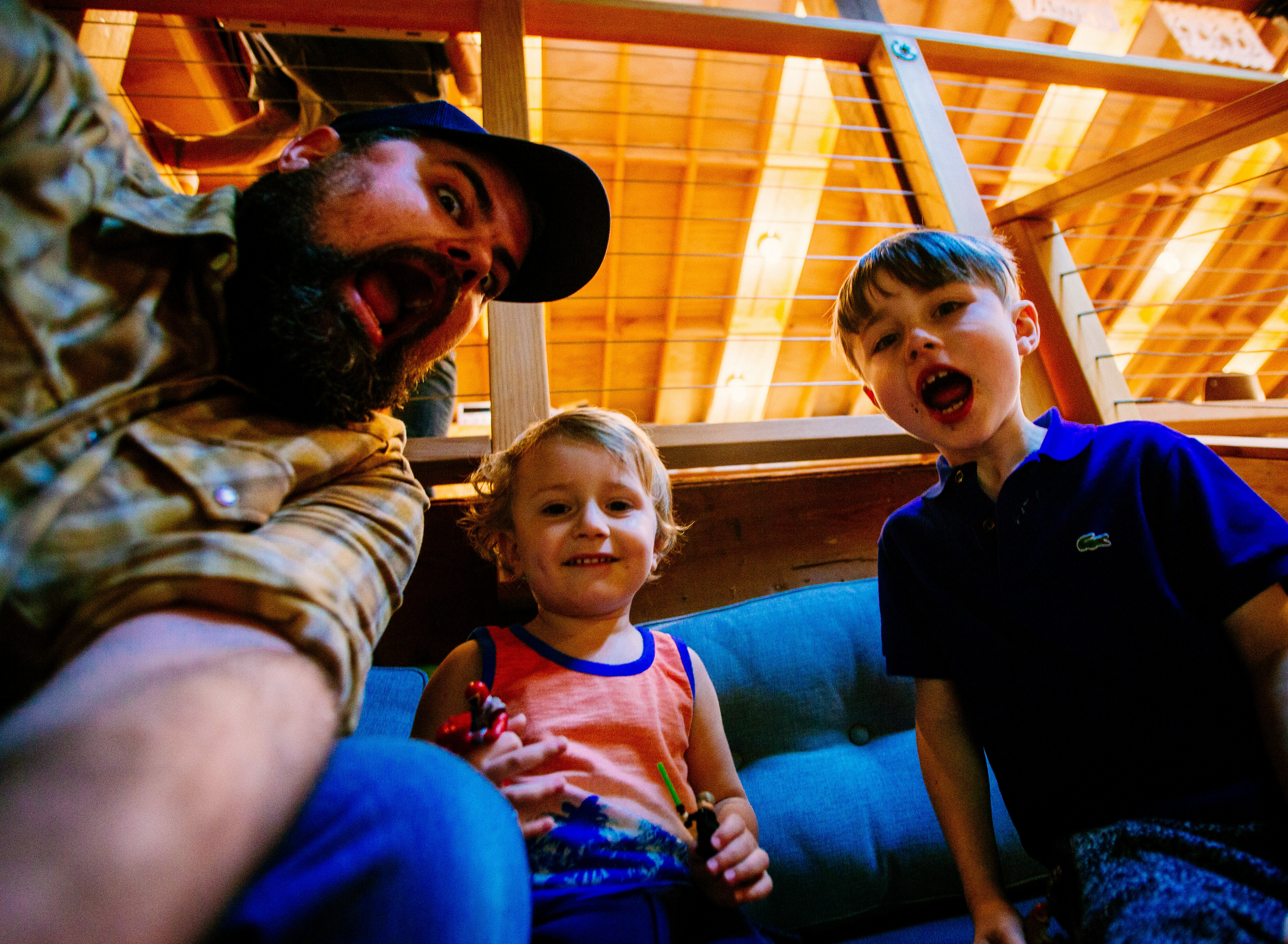 Bearded man in a cap taking a selfie with two young kids inside a wooden cabin