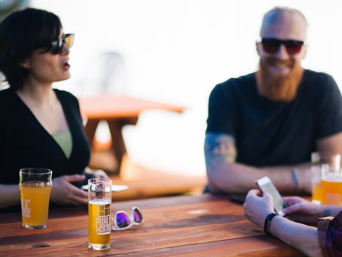 Friends in sunglasses laughing over Public House beer flights at an outdoor table