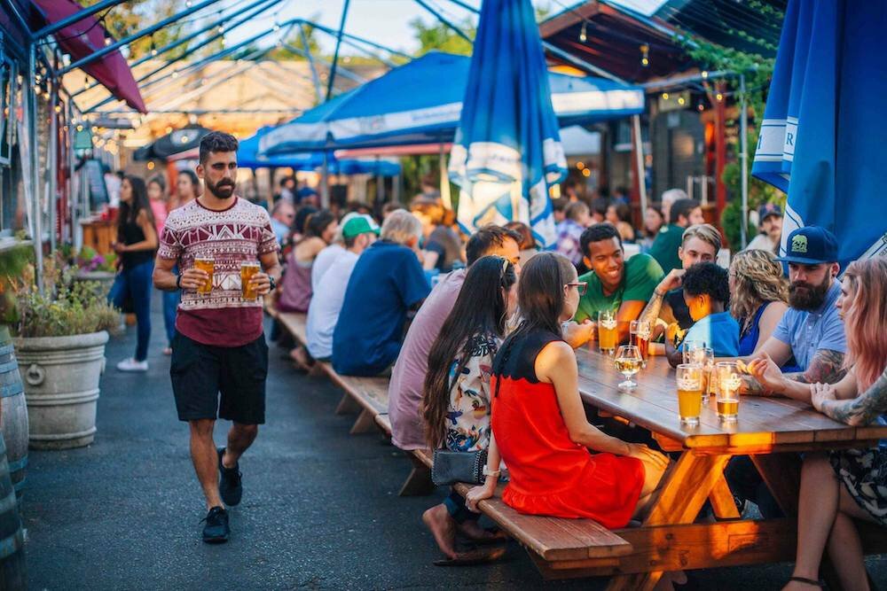 Man carrying a beer past long communal tables of patrons at an outdoor beer garden in Eugene