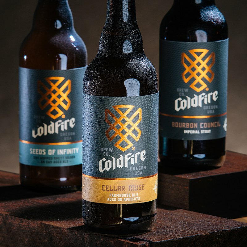 Trio of ColdFire Brewing bottles including Cellar Muse farmhouse ale on dark wood blocks