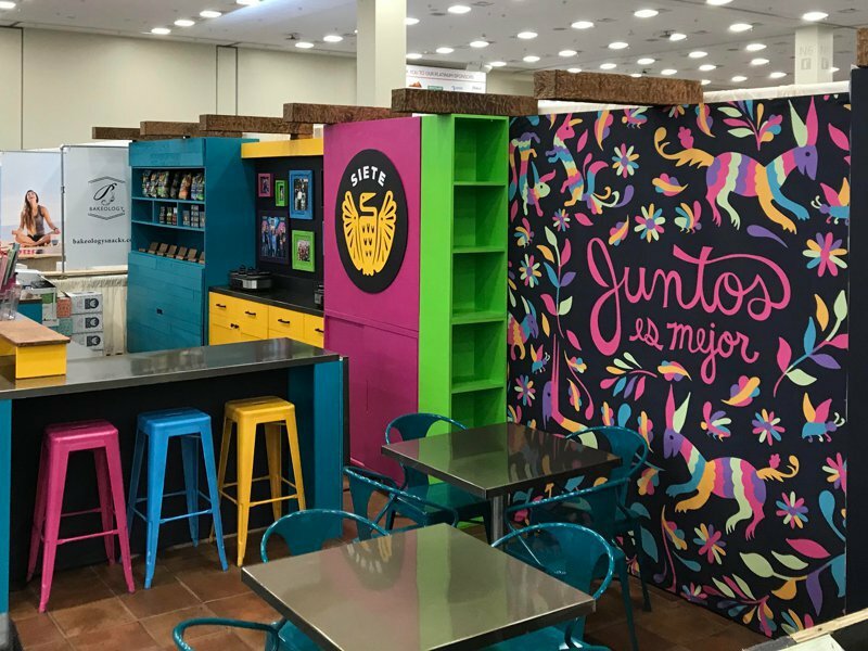 Siete trade show booth with colorful Otomi mural reading Juntos es Mejor and neon stools