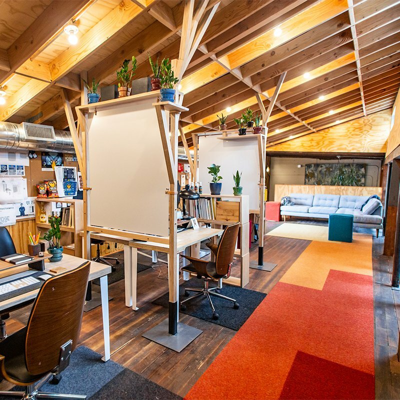 Drawn's Bard Building studio loft with wood-beam ceiling, drafting desks topped by potted plants, and orange-toned carpet