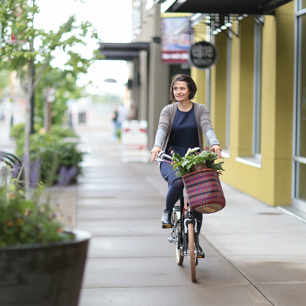 Woman riding a bicycle with a flower-filled basket along a downtown Eugene sidewalk