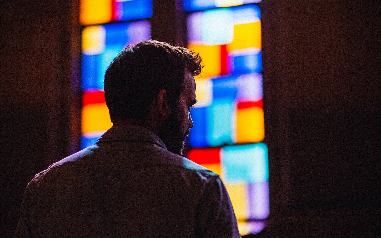 Bearded man seen from behind gazing at a colorful stained-glass window in a dim space