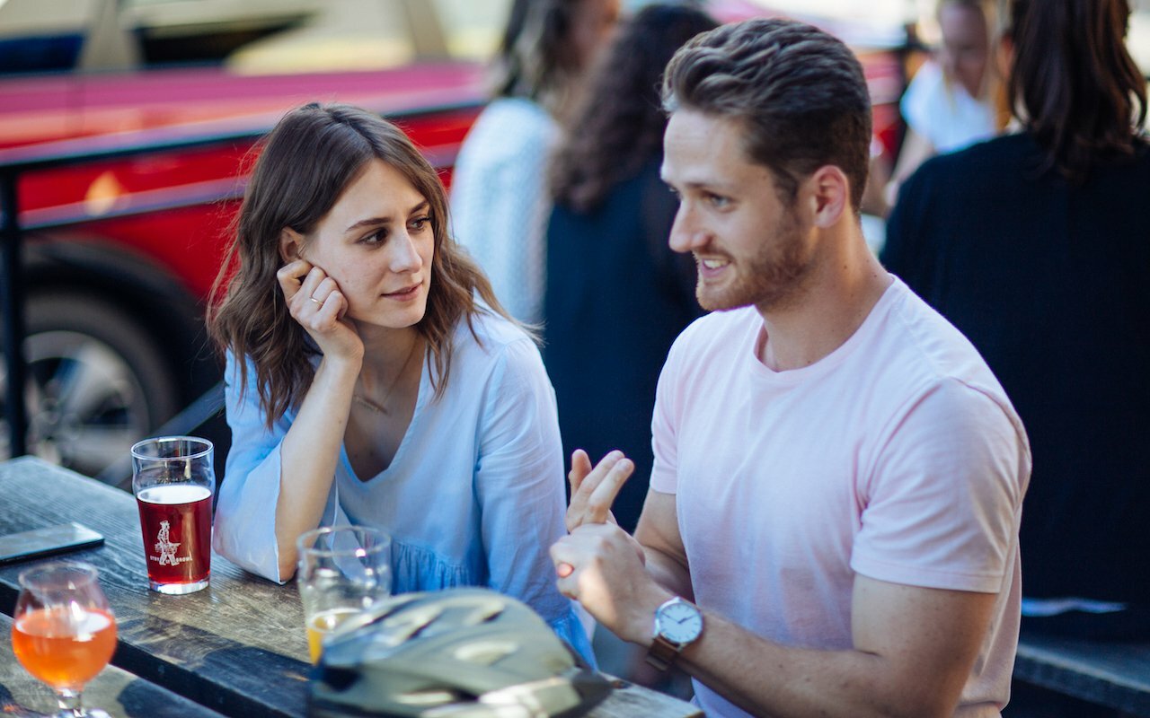 Man and woman talking over pints at an outdoor table, man gesturing mid-conversation