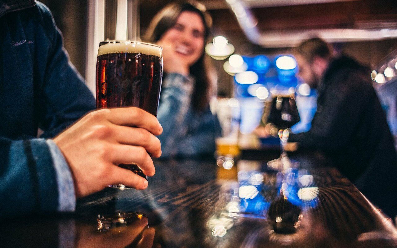 Hand holding a glass of dark beer at a bar as a woman laughs in the background