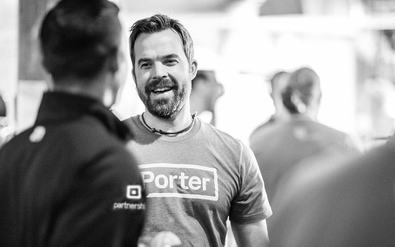 Smiling man in a Porter partnership tee talking with people at an outdoor event, black-and-white