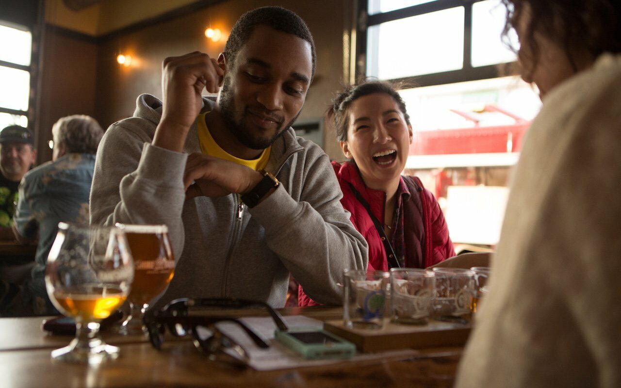 Three friends laughing and sharing beers at a wood pub table, growlers and glasses in the foreground