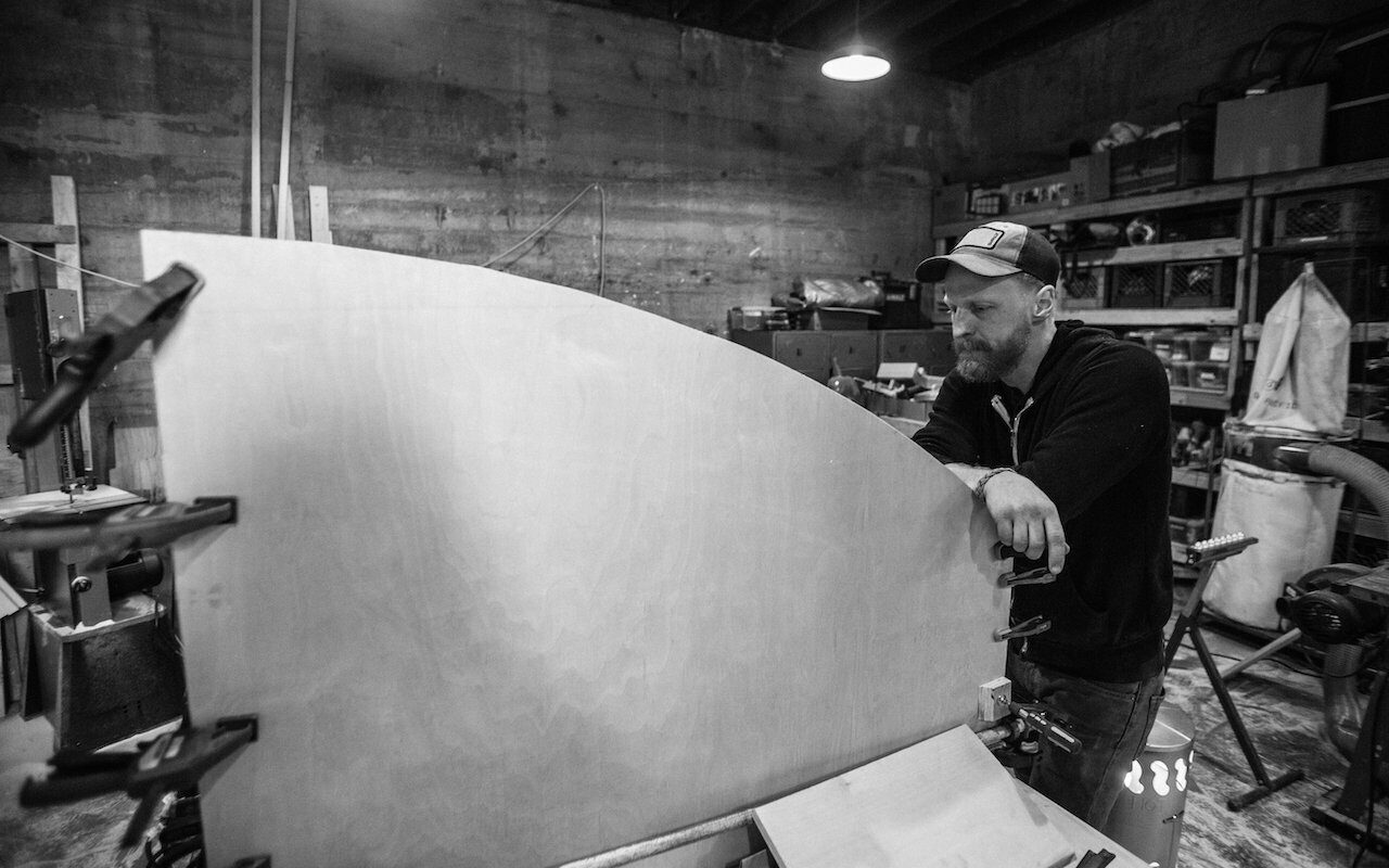 Man in a cap leaning over a large curved fabricated panel in a workshop, black-and-white