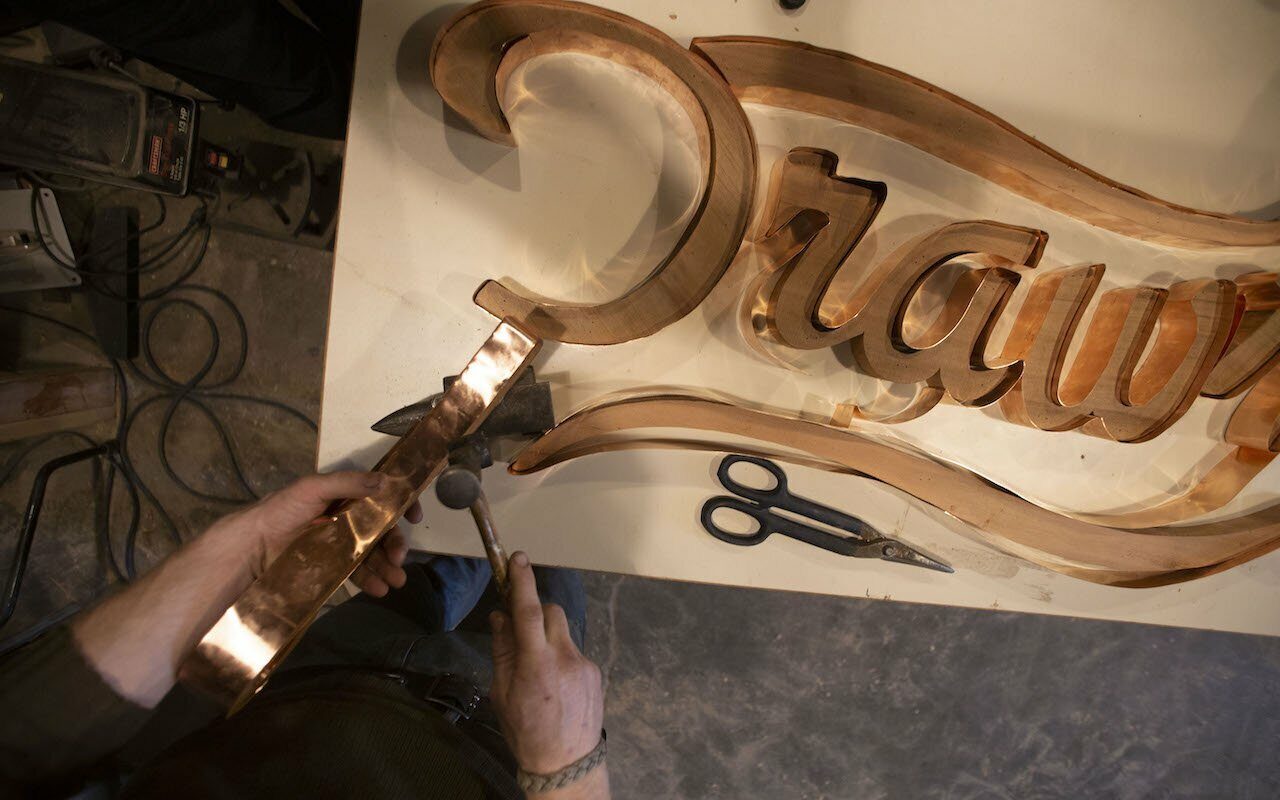 Overhead view of hands hammering copper around a carved wooden 'Drawn' script sign in a shop