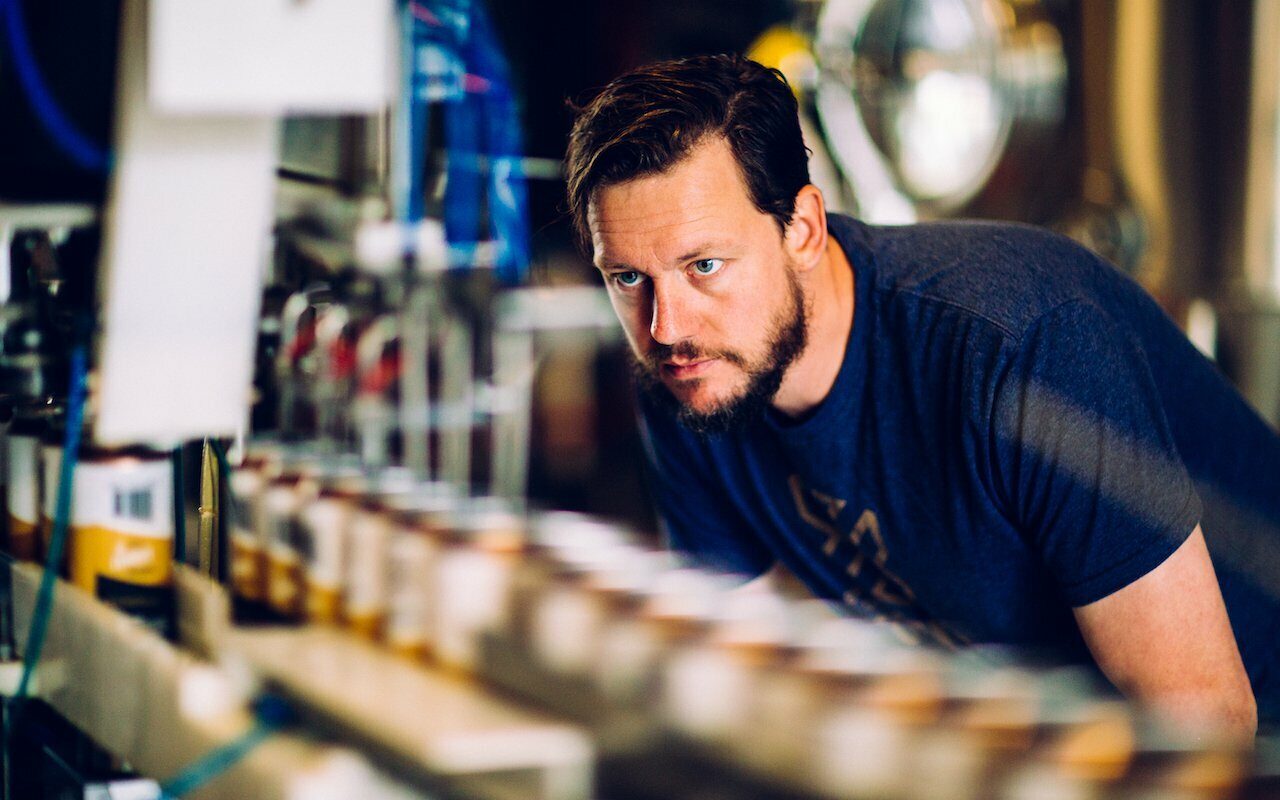 Focused man in a navy tee beside a canning line of beer cans at Coldfire brewery