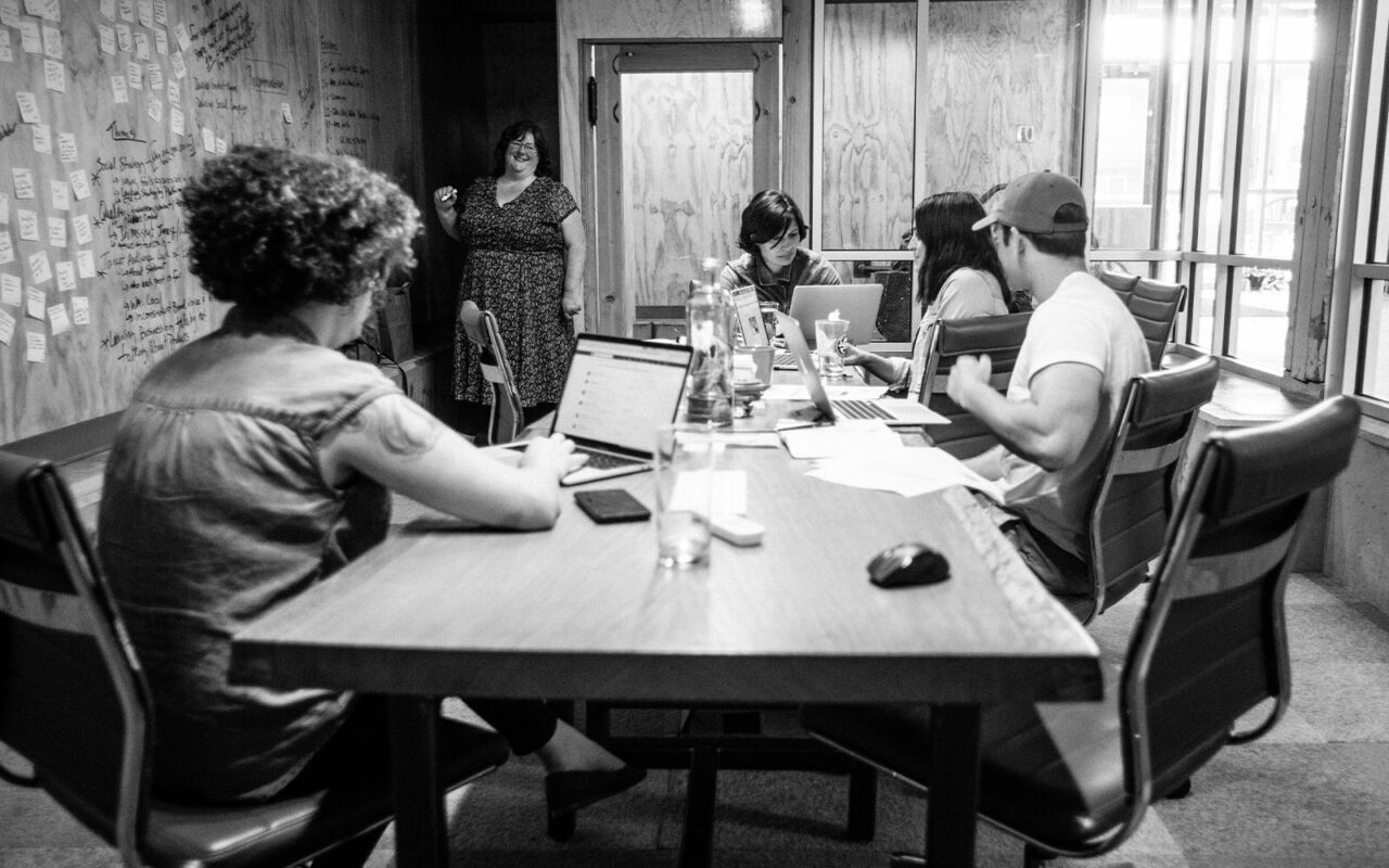 Drawn team in a black-and-white brand audit workshop, gathered at a table by a wall of sticky notes