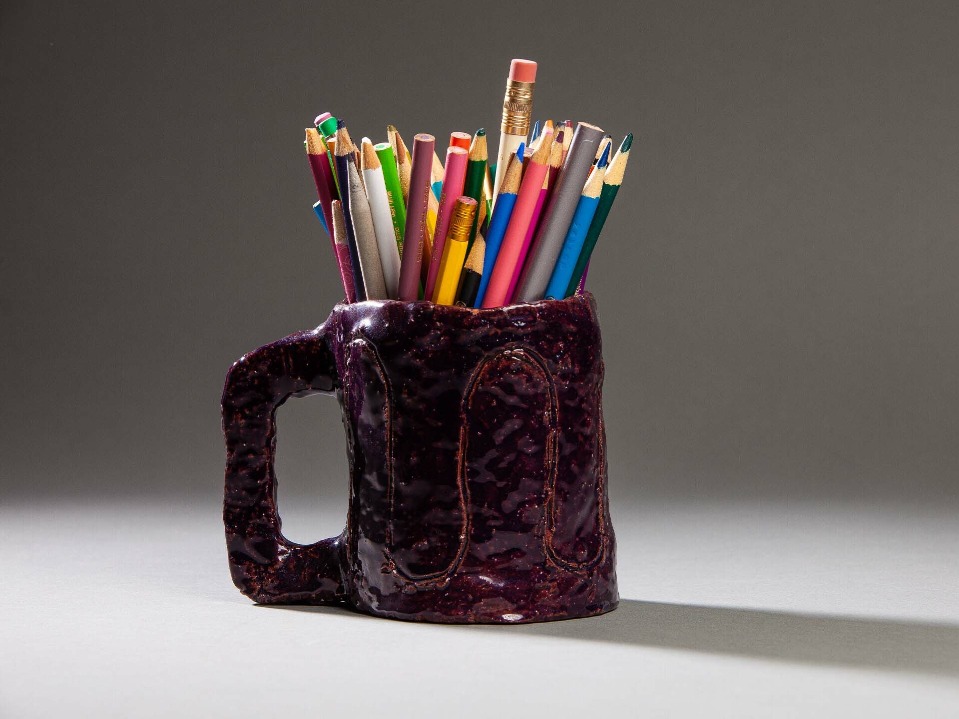 Lumpy handmade purple ceramic mug filled with colored pencils, side view