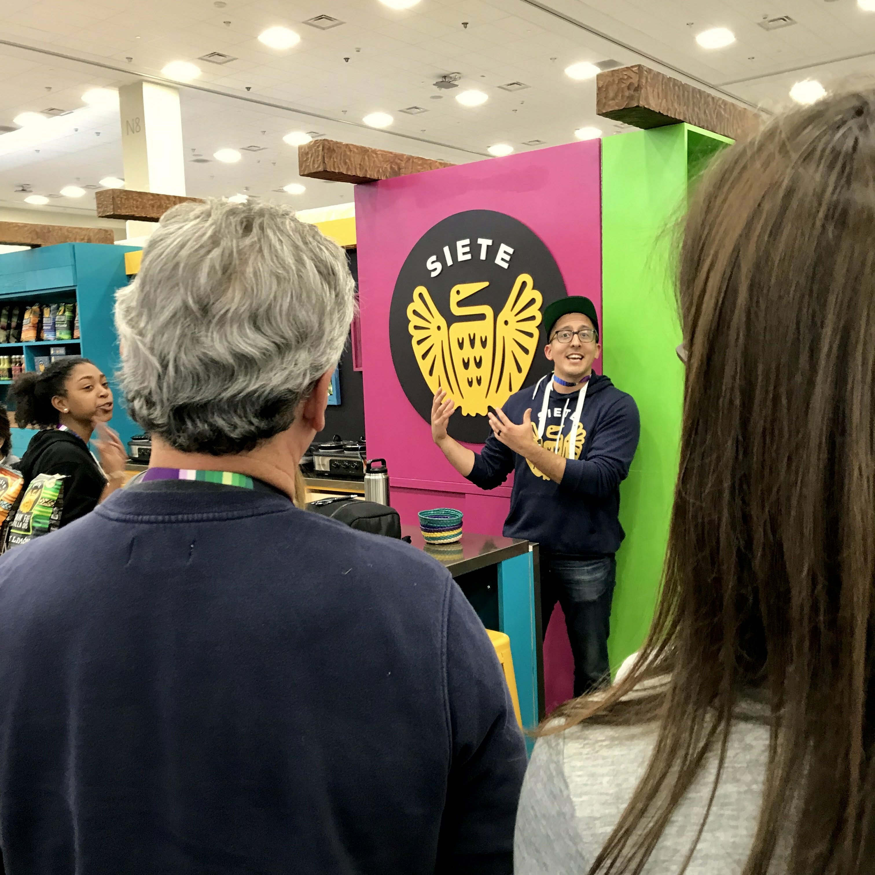 Presenter speaking by the Siete logo wall as visitors watch at the trade show booth