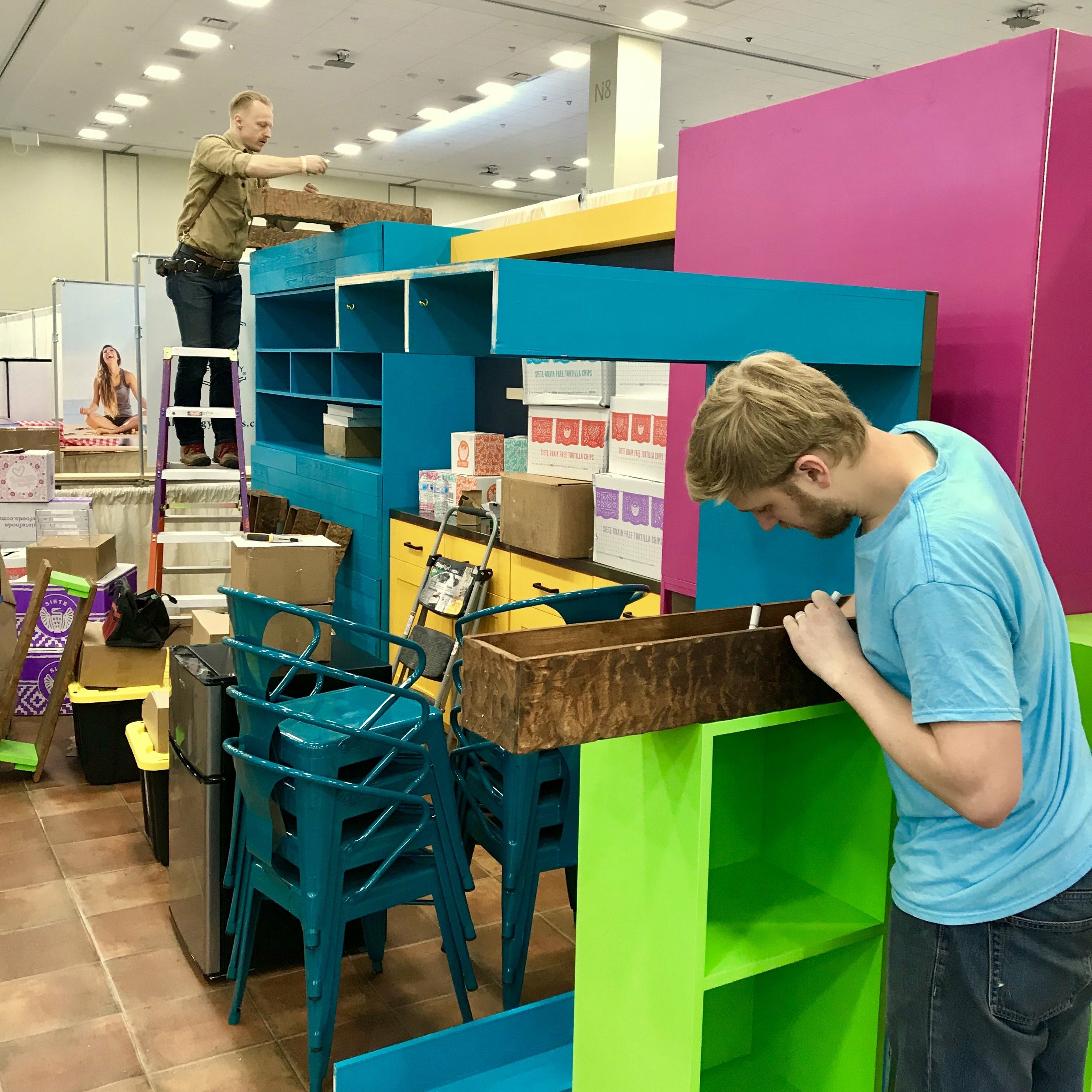 Crew assembling Siete's colorful trade show booth shelving amid shipping boxes