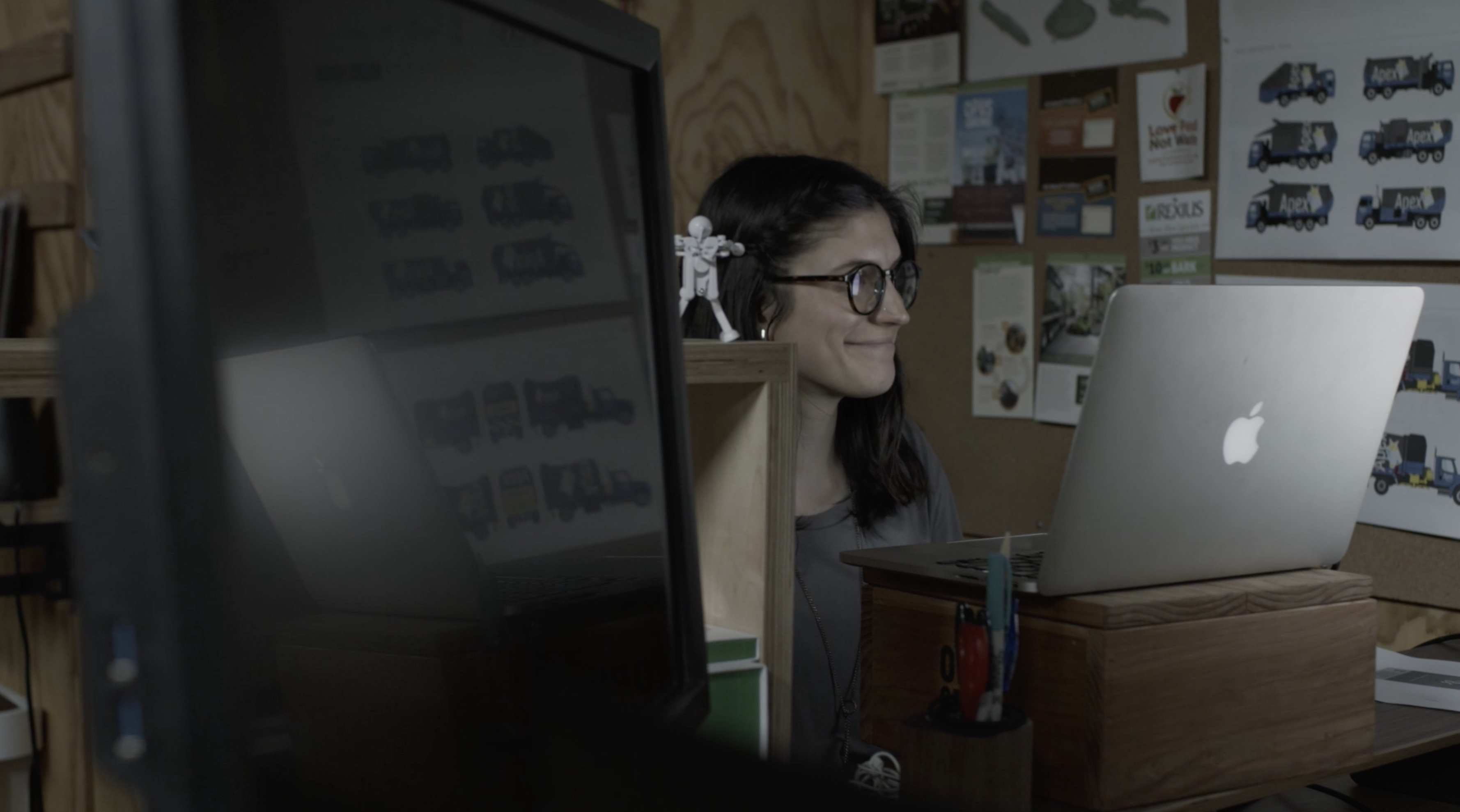Woman in glasses smiling at a laptop and monitor in a workshop studio