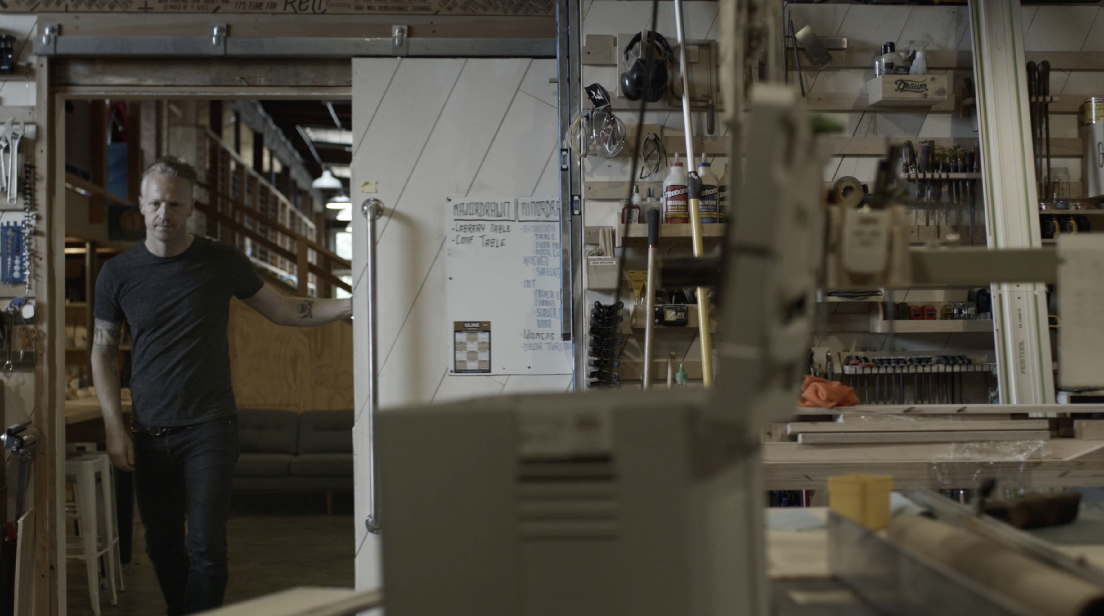Man standing in a Drawn fabrication workshop beside a whiteboard and lumber racks