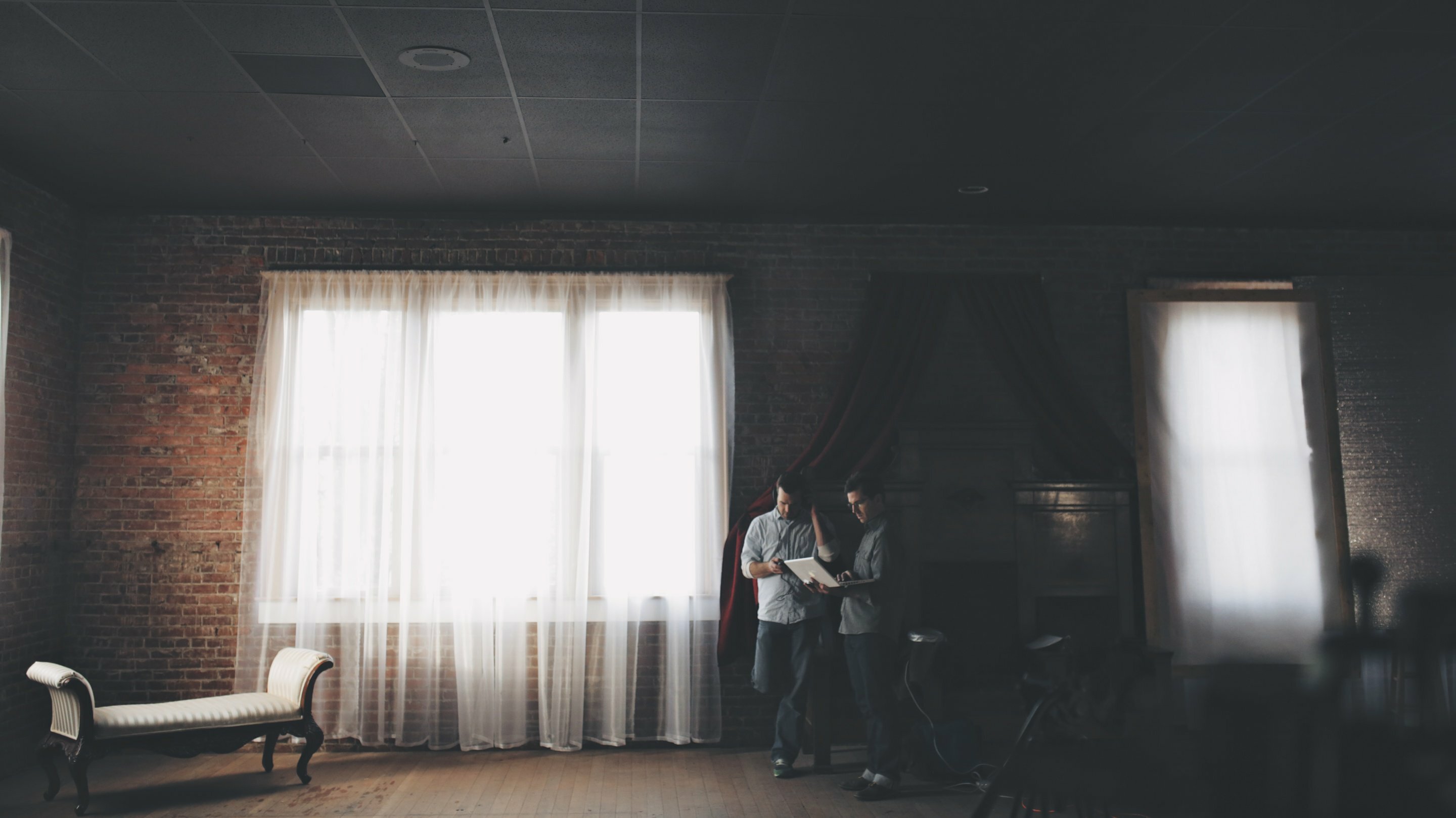 Two men reviewing plans by a sunlit curtained window in a dim brick-walled room