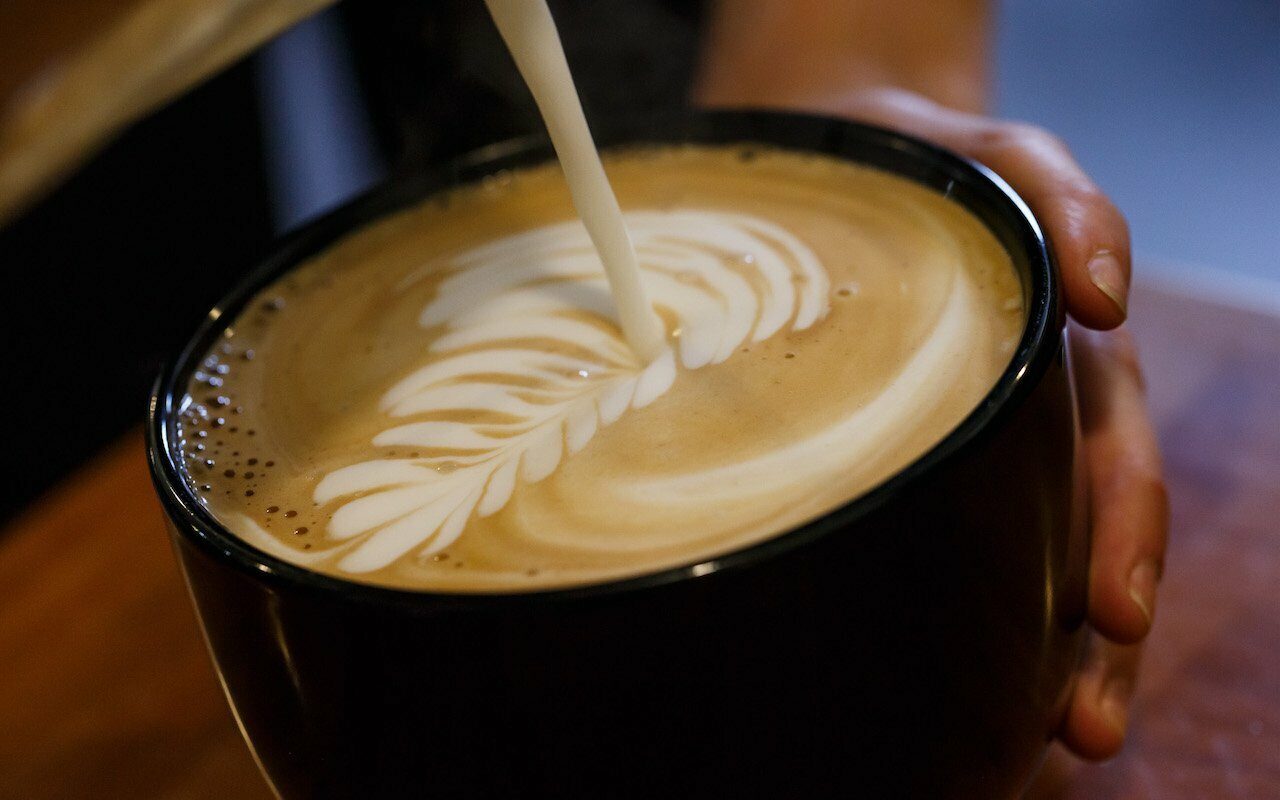 Barista pouring milk to make leaf latte art in a dark cup held in hand