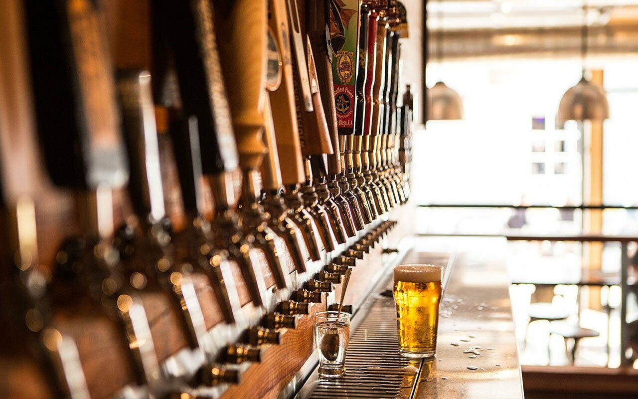 Long row of brewery beer taps with a full glass of golden beer by a window