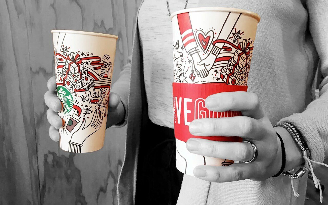 Hands holding two illustrated Starbucks holiday Give Good cups, one in a red sleeve, partly desaturated