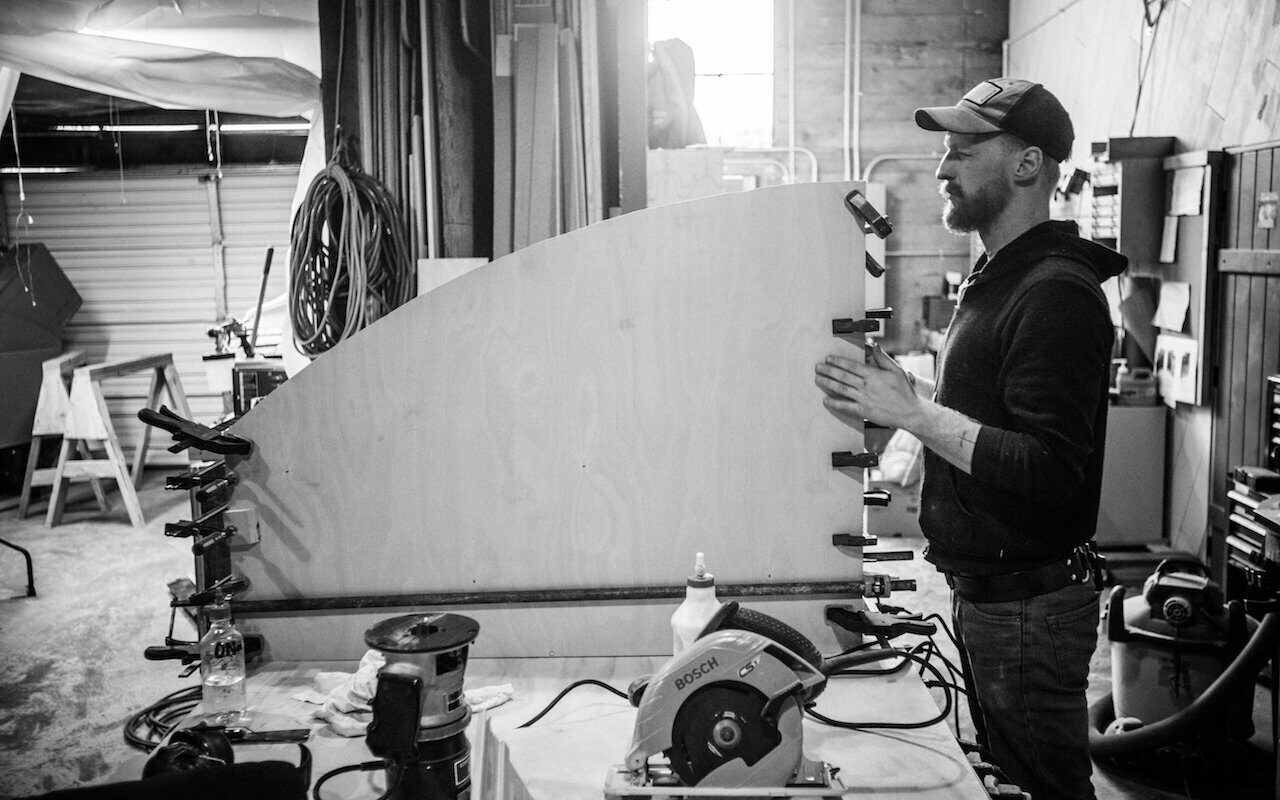 Maker guiding a curved plywood form past a Bosch circular saw in the fabrication workshop, black and white
