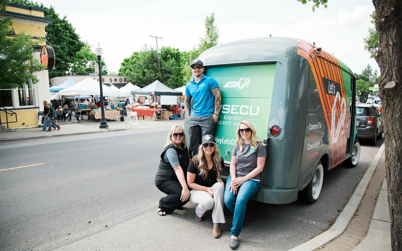 Four people posing with a branded WSECU credit union trailer at a street market