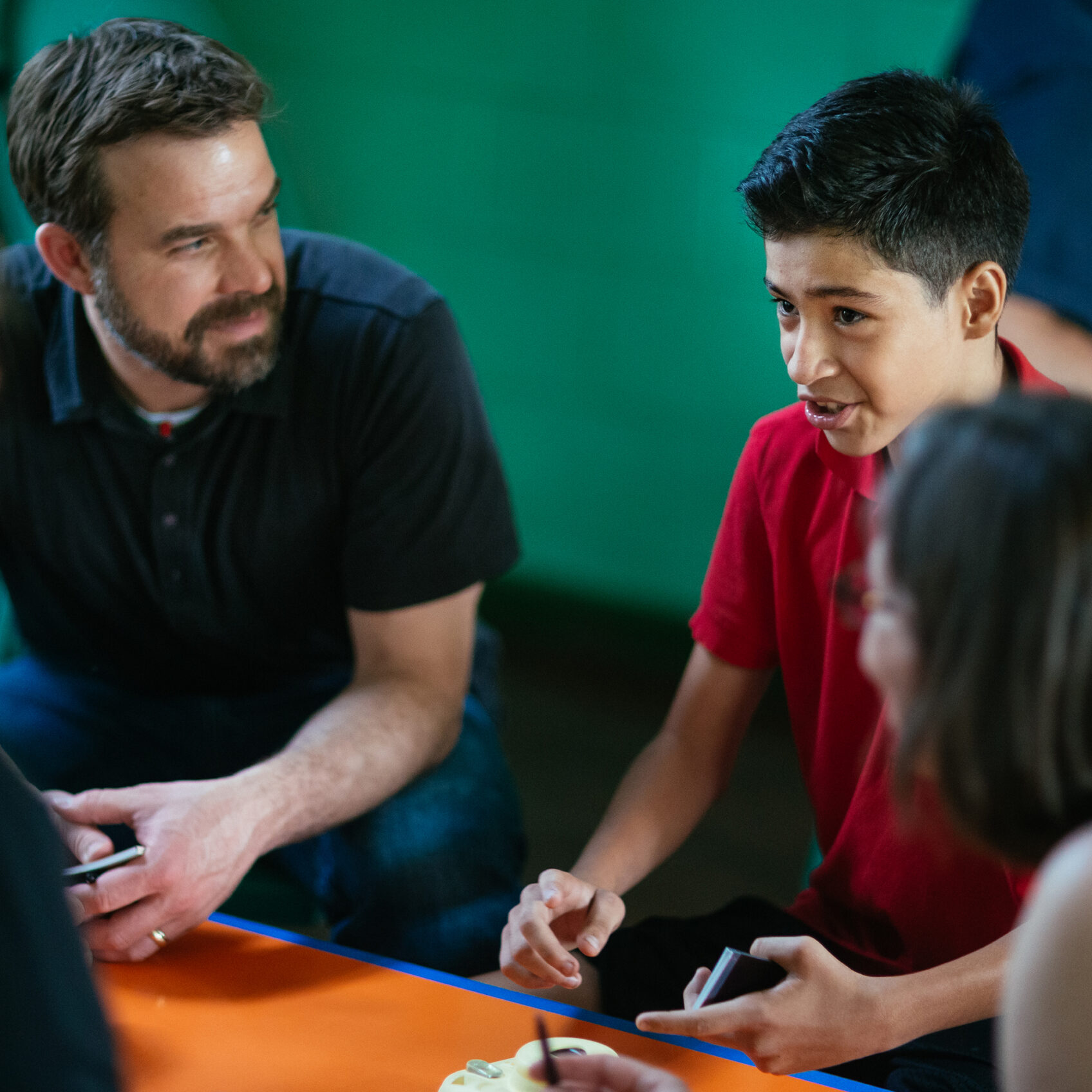 Bearded facilitator talking with kids around an orange table during a workshop