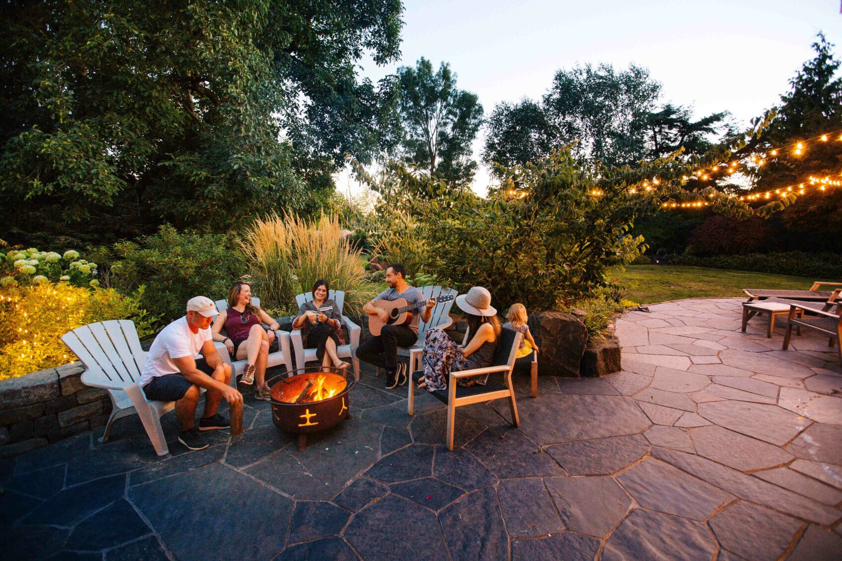 Group relaxing in chairs around a backyard fire pit under string lights at dusk