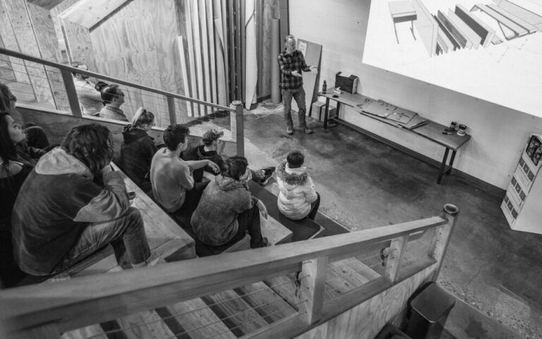 Apprentices seated on steps watching an instructor present at a design fabrication shop, black-and-white