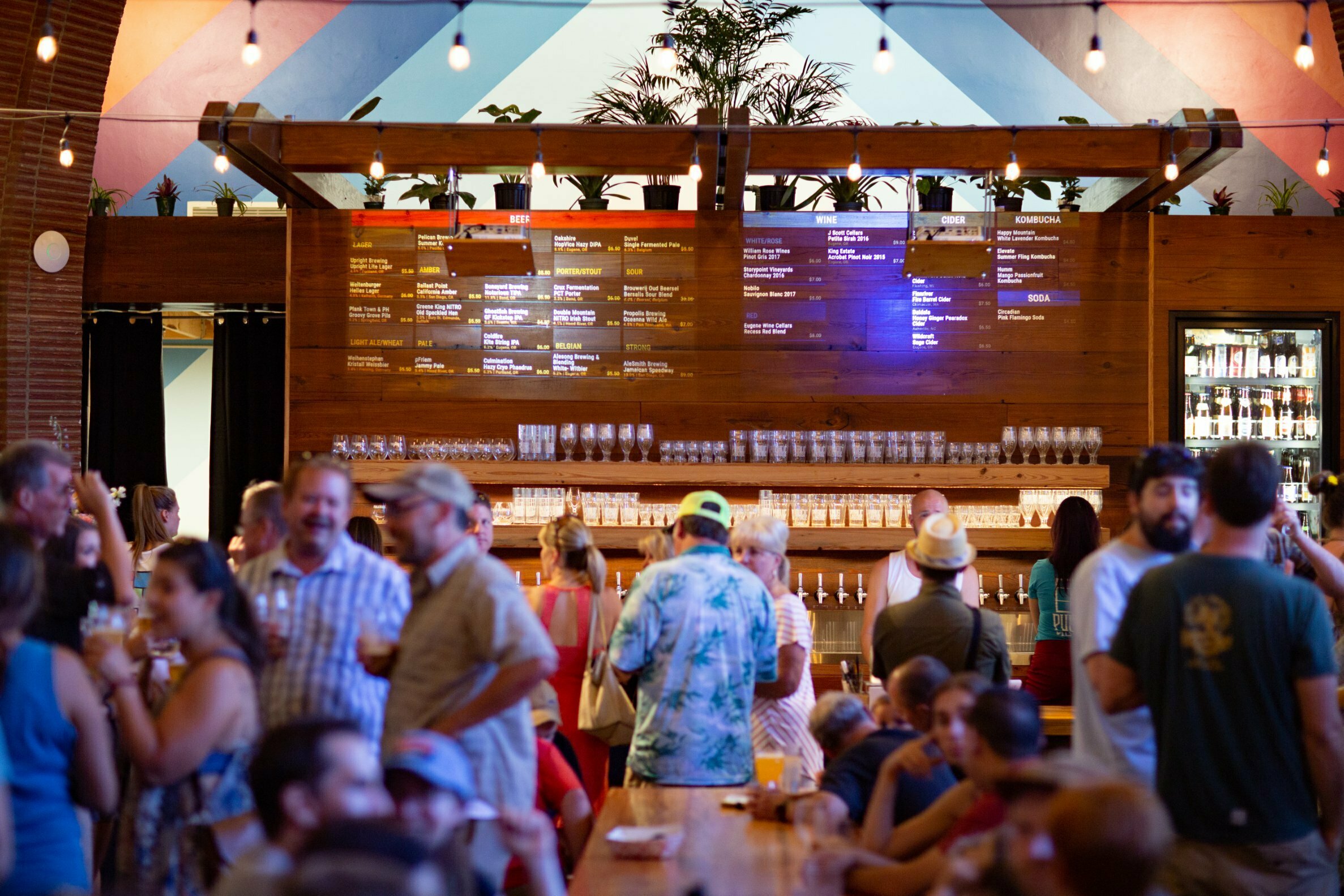 Crowd gathering at PublicHouse food hall bar beneath illuminated menu boards and string lights