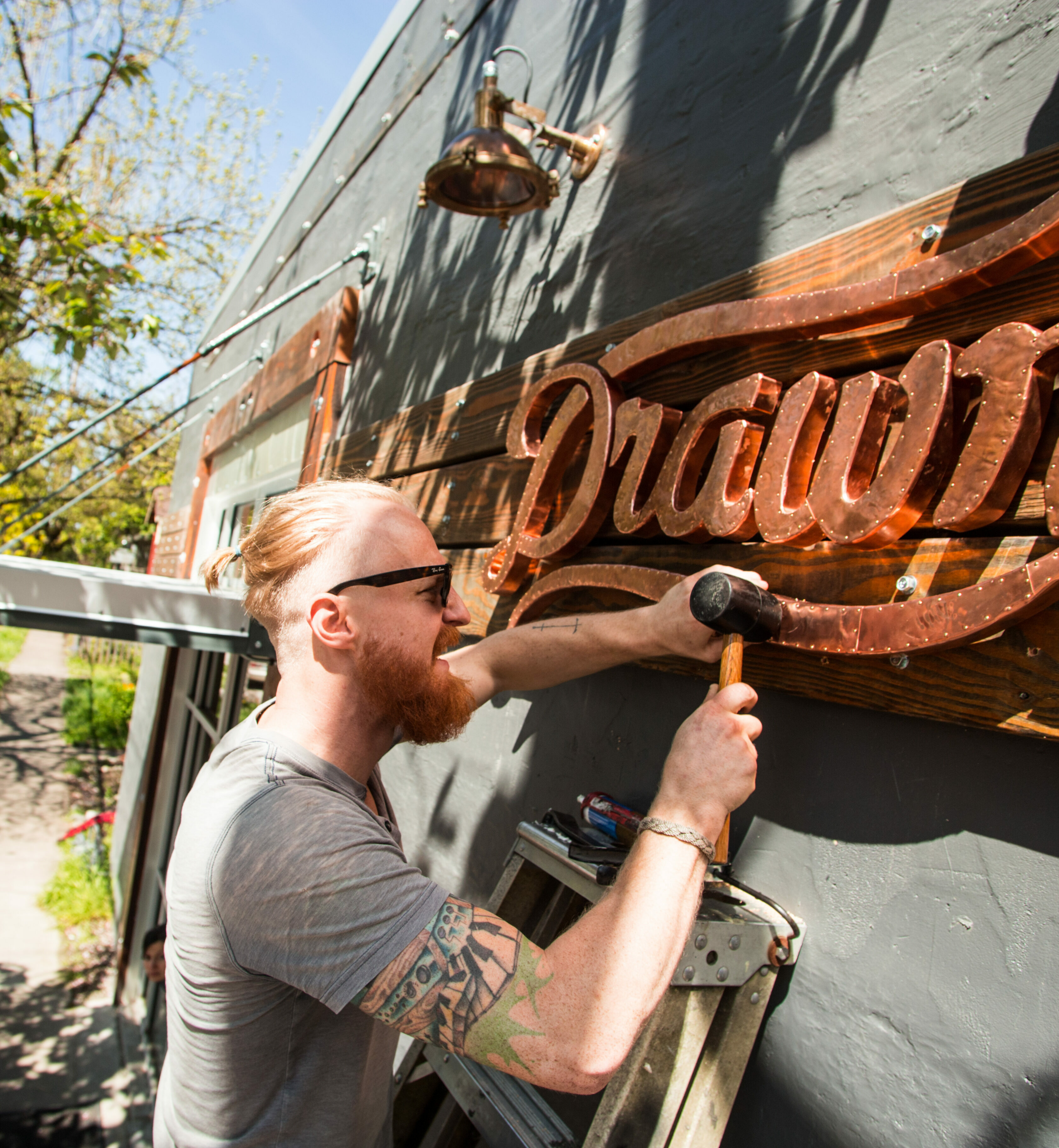 Man hammering a carved wooden Drawn sign onto a gray studio wall outdoors
