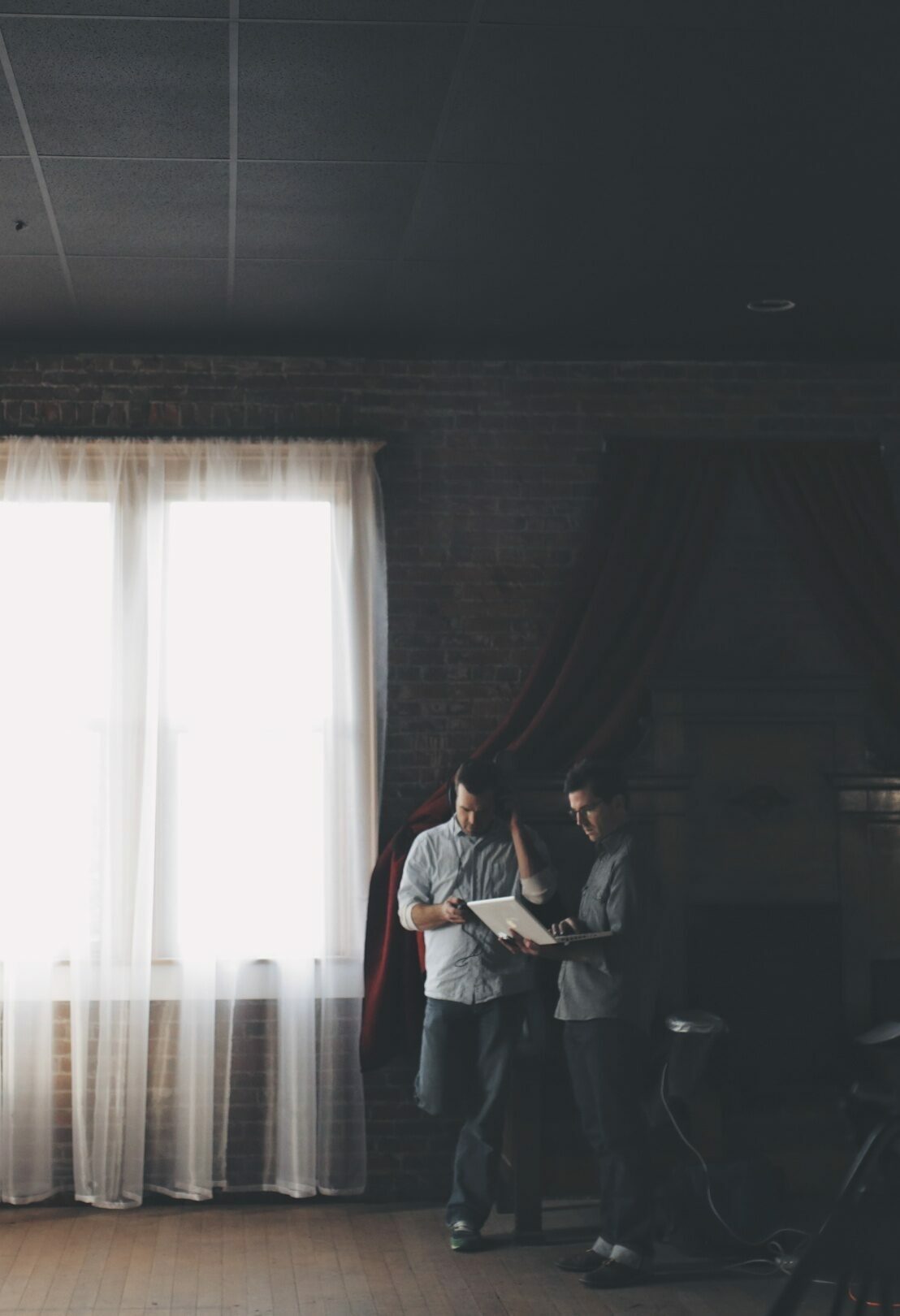 Two men reviewing a document by a bright curtained window in a dim room