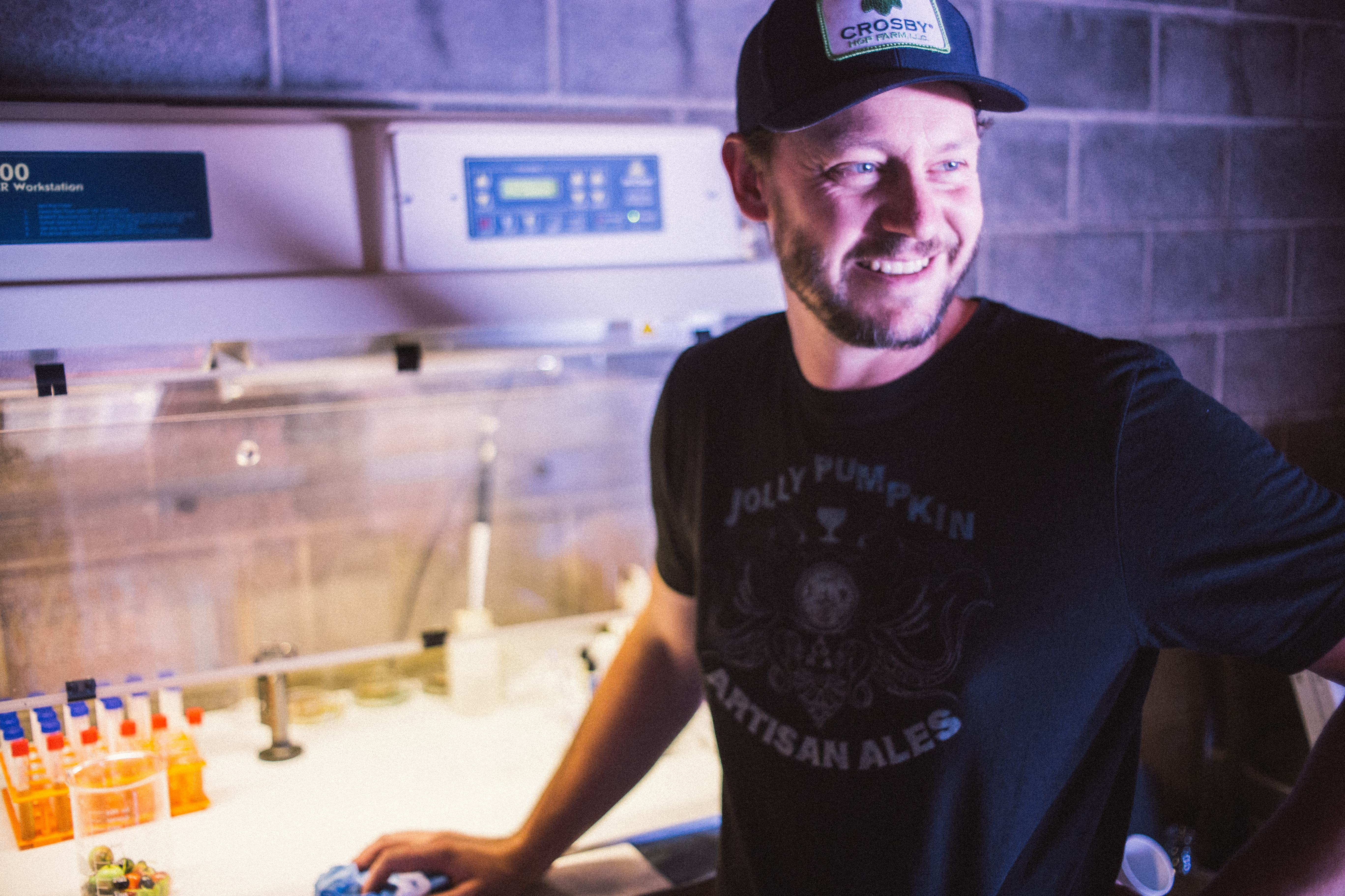 Smiling man in a Jolly Pumpkin Artisan Ales tee and Crosby cap working in a Coldfire brewery lab
