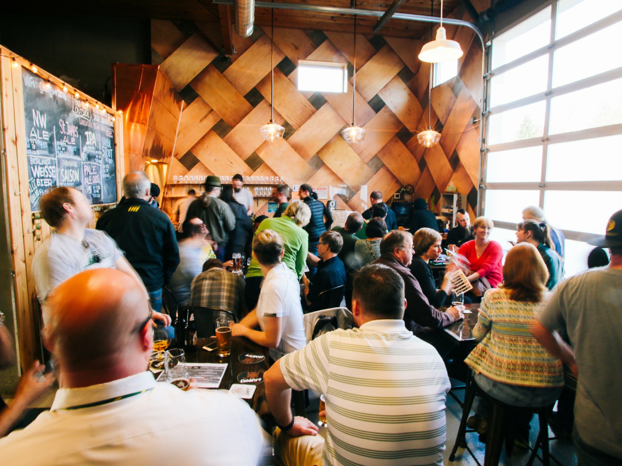 Crowded taproom with people seated at tables before a woven wood feature wall and chalkboard beer menu