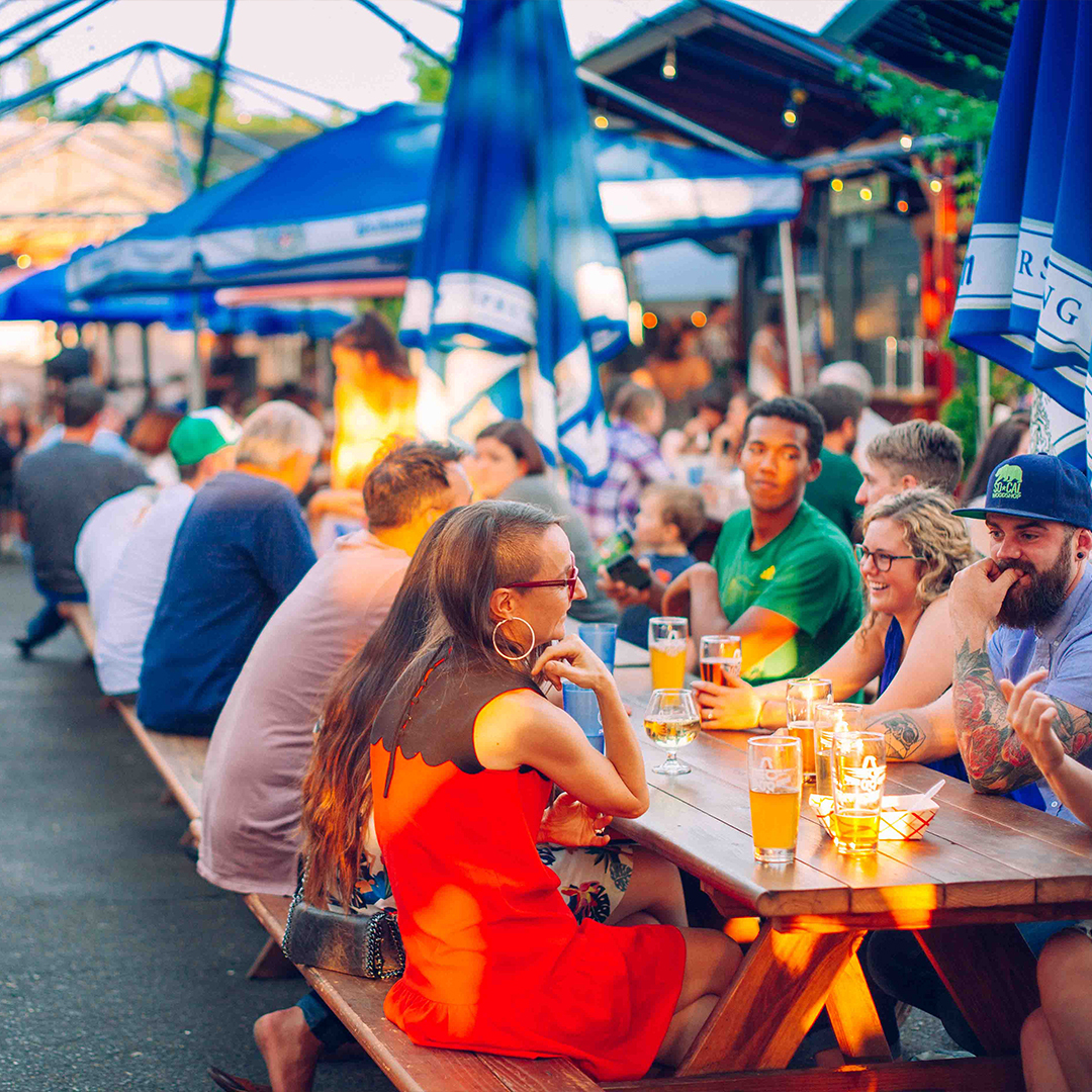 Patrons in summer clothes drinking beer at long picnic tables under blue umbrellas at an outdoor beer garden